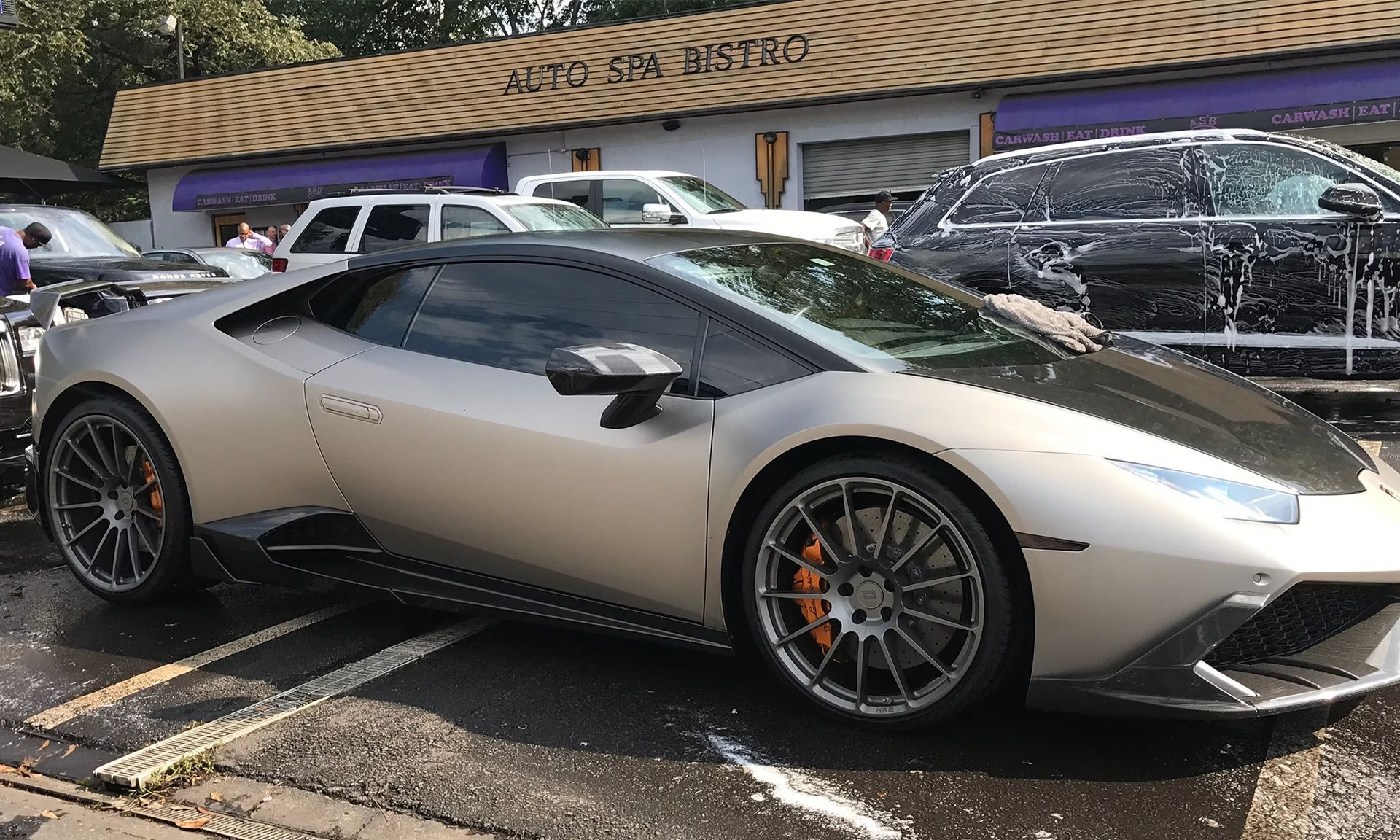 A matte champagne-colored Lamborghini Huracán parked in a lot in front of a building with a brown awning.