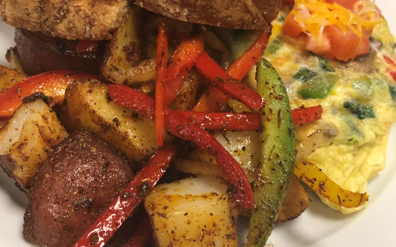A plate of breakfast featuring home fries with sliced red and green peppers, served alongside a vegetable omelet.