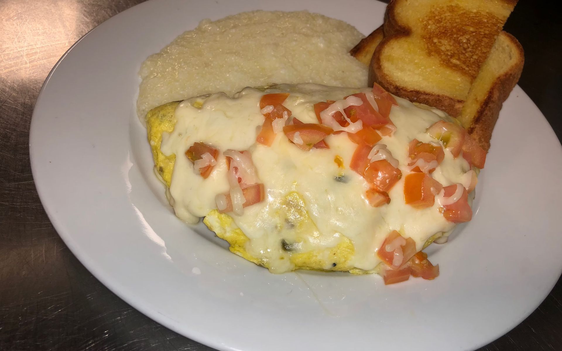 Omelet topped with melted cheese and diced tomatoes, with grits and toast on a white plate.