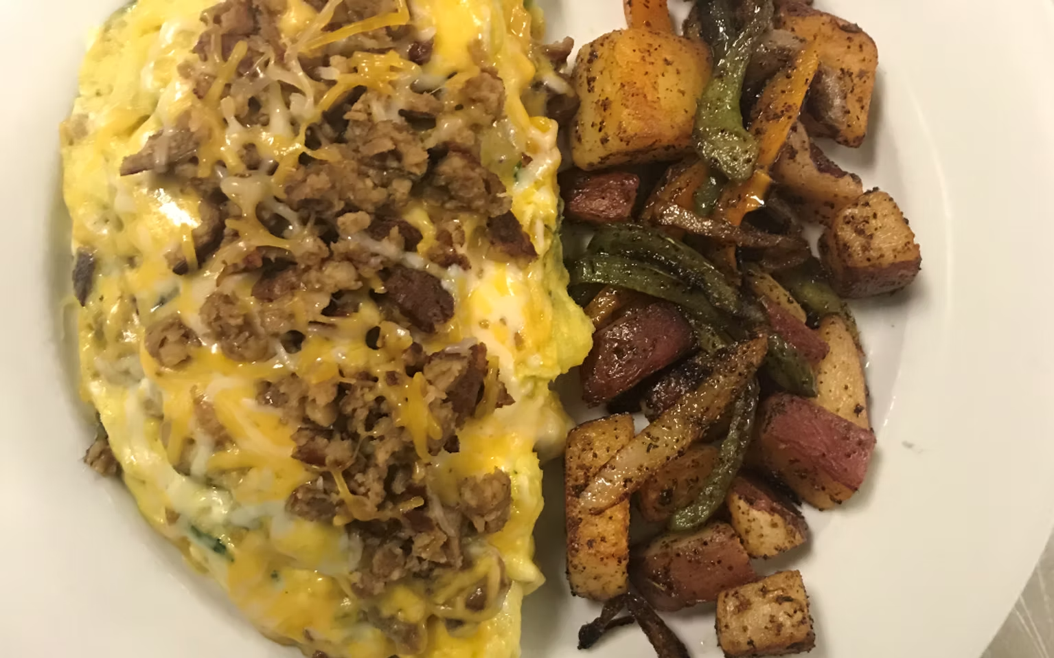 Omelet with sausage and cheese, next to roasted potatoes, red onion, and green peppers on a white plate.