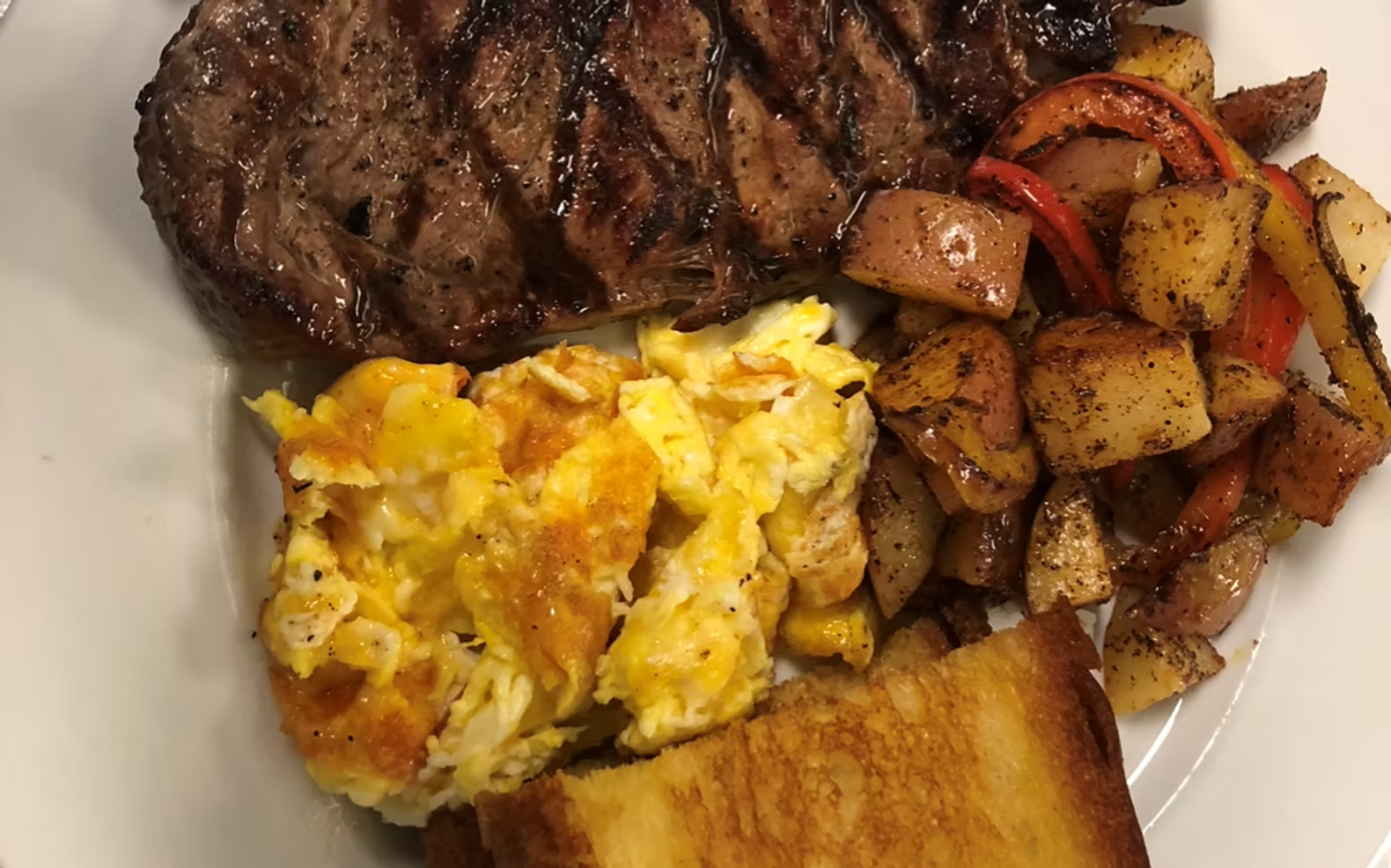 Grilled steak with scrambled eggs, potatoes, and toast on a plate.