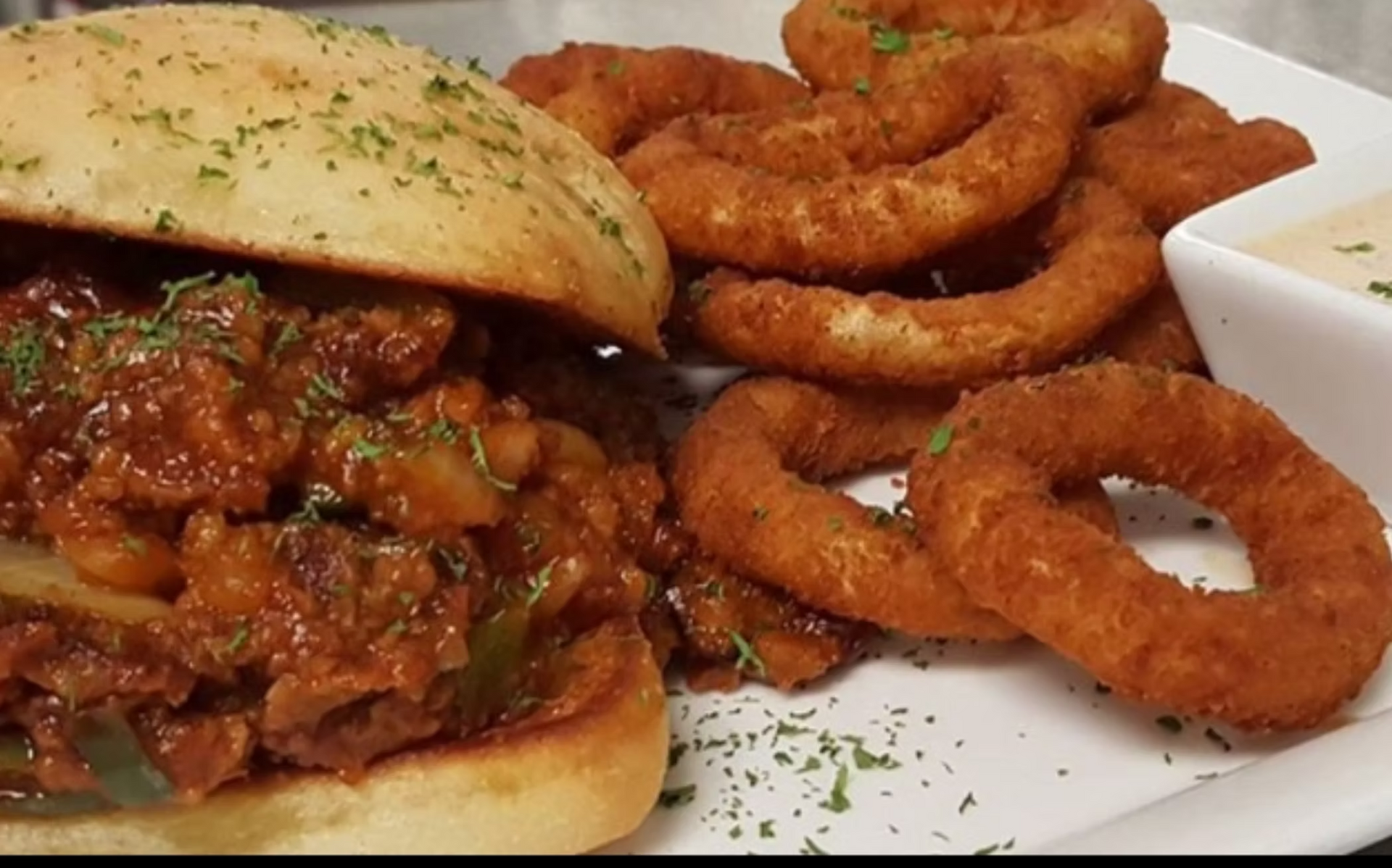 Sloppy joe sandwich with onion rings and dipping sauce on a white plate.