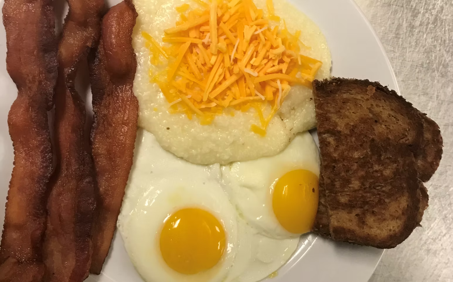 Breakfast plate: bacon, grits with shredded cheese, two sunny-side-up eggs, and toasted bread.