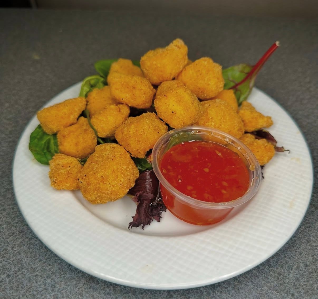A pile of golden-brown fried nuggets served on a bed of fresh greens with a side of red dipping sauce on a white plate.