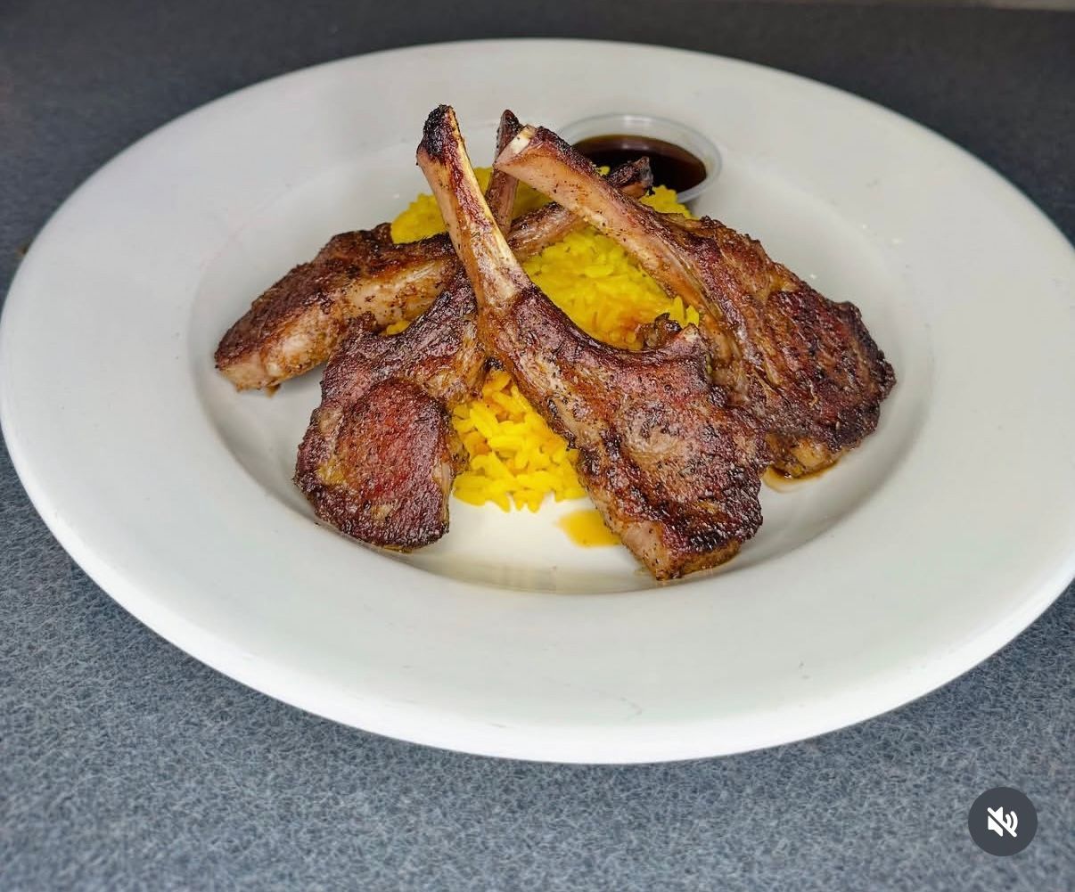 Three grilled lamb chops served over a mound of bright yellow saffron rice, with a small ramekin of dark dipping sauce.