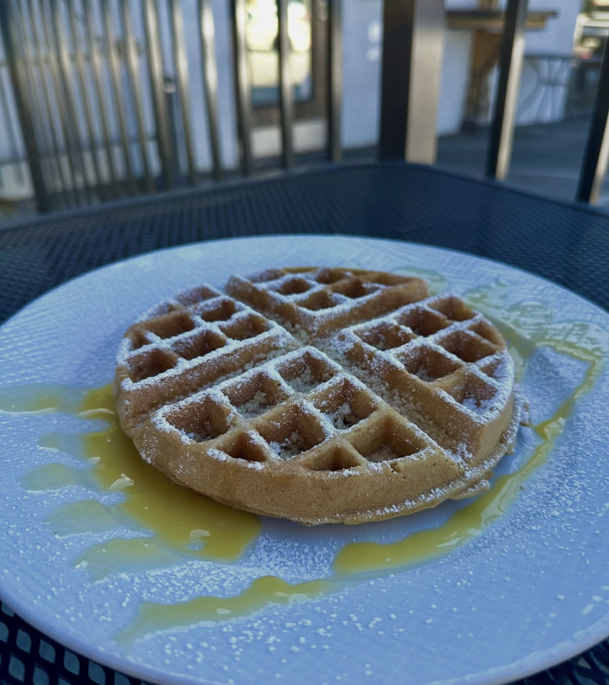 A round waffle dusted with powdered sugar and drizzled with yellow syrup on a white plate on a dark outdoor table.