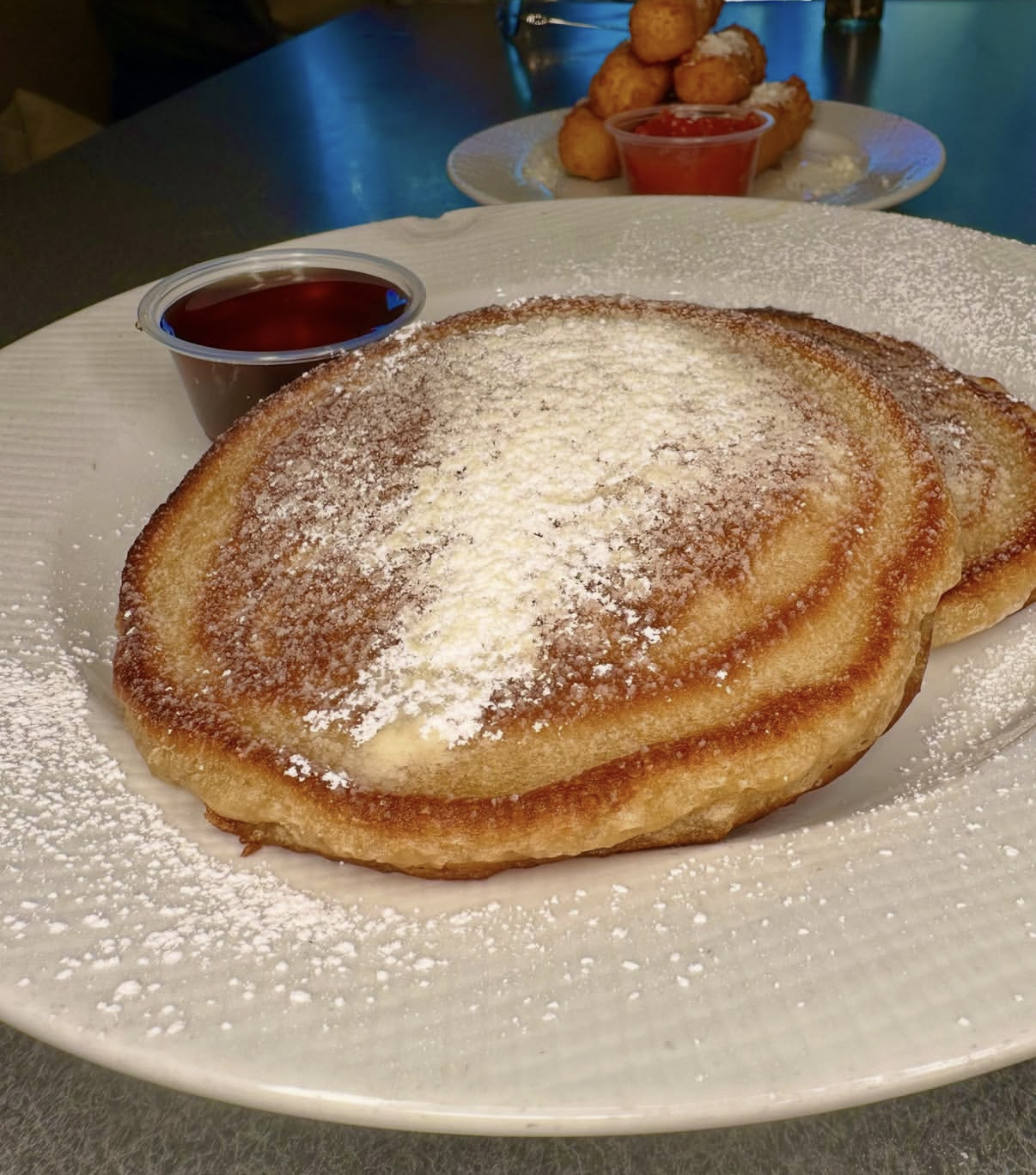 Two pancakes dusted with powdered sugar on a white plate, served with a side of syrup and a plate of fried dough.