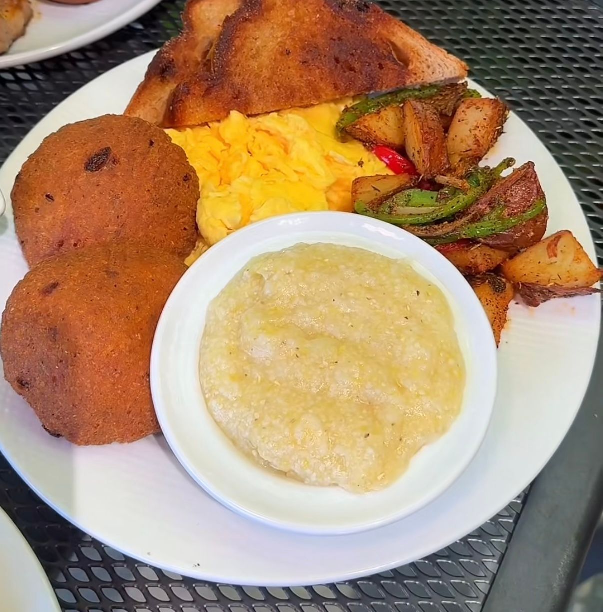 A breakfast plate with two fried hushpuppies, scrambled eggs, toasted bread, sautéed potatoes, and a bowl of grits.