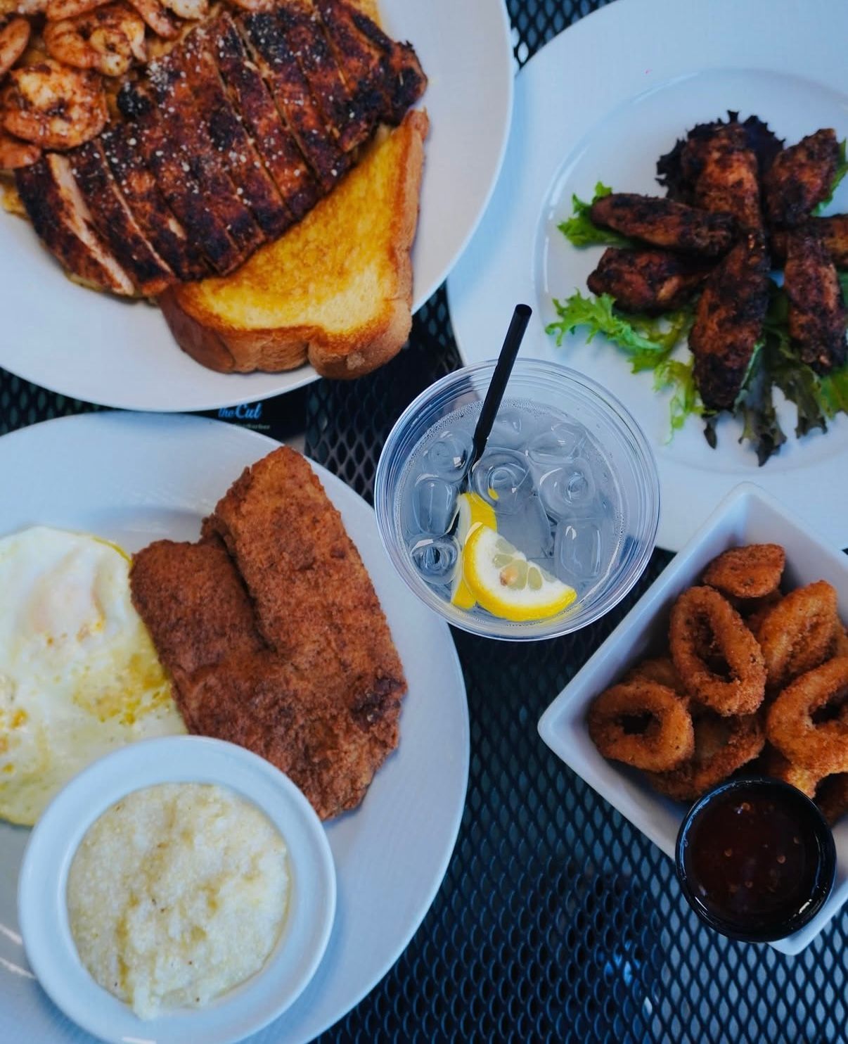 A top-down view of a table spread with several plates of food, including fried fish, grits, steak, and onion rings.