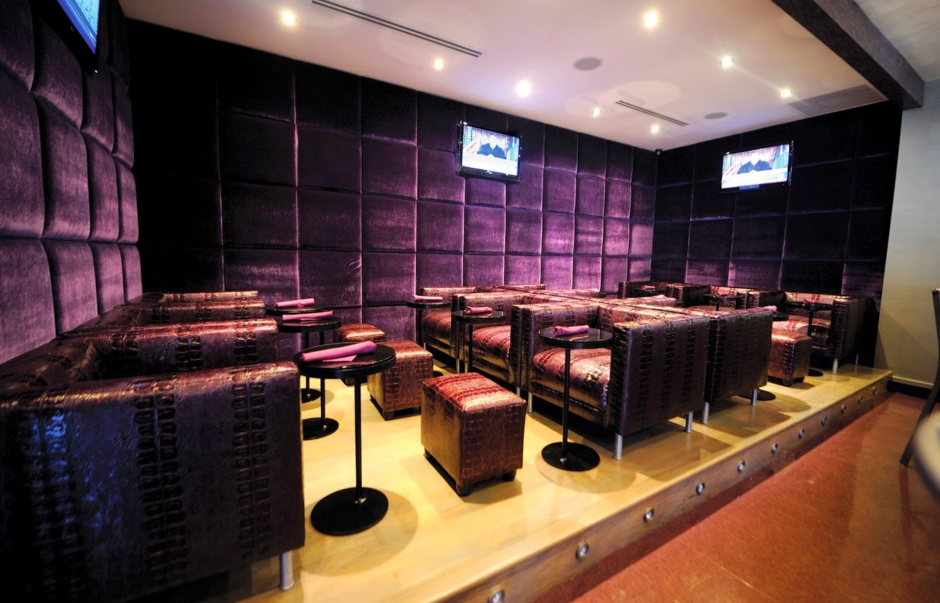 A dimly lit lounge featuring rows of dark leather seats, small tables, and purple padded walls with mounted screens.