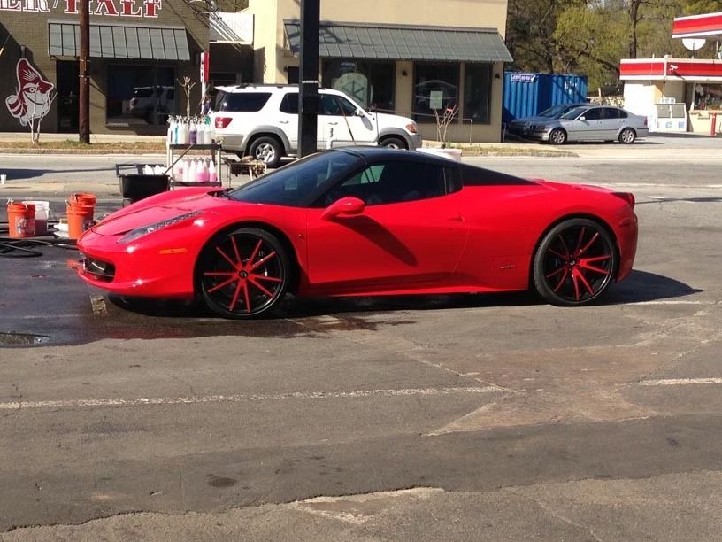 A red Ferrari sports car with custom red and black wheels parked in an outdoor lot on a sunny day.