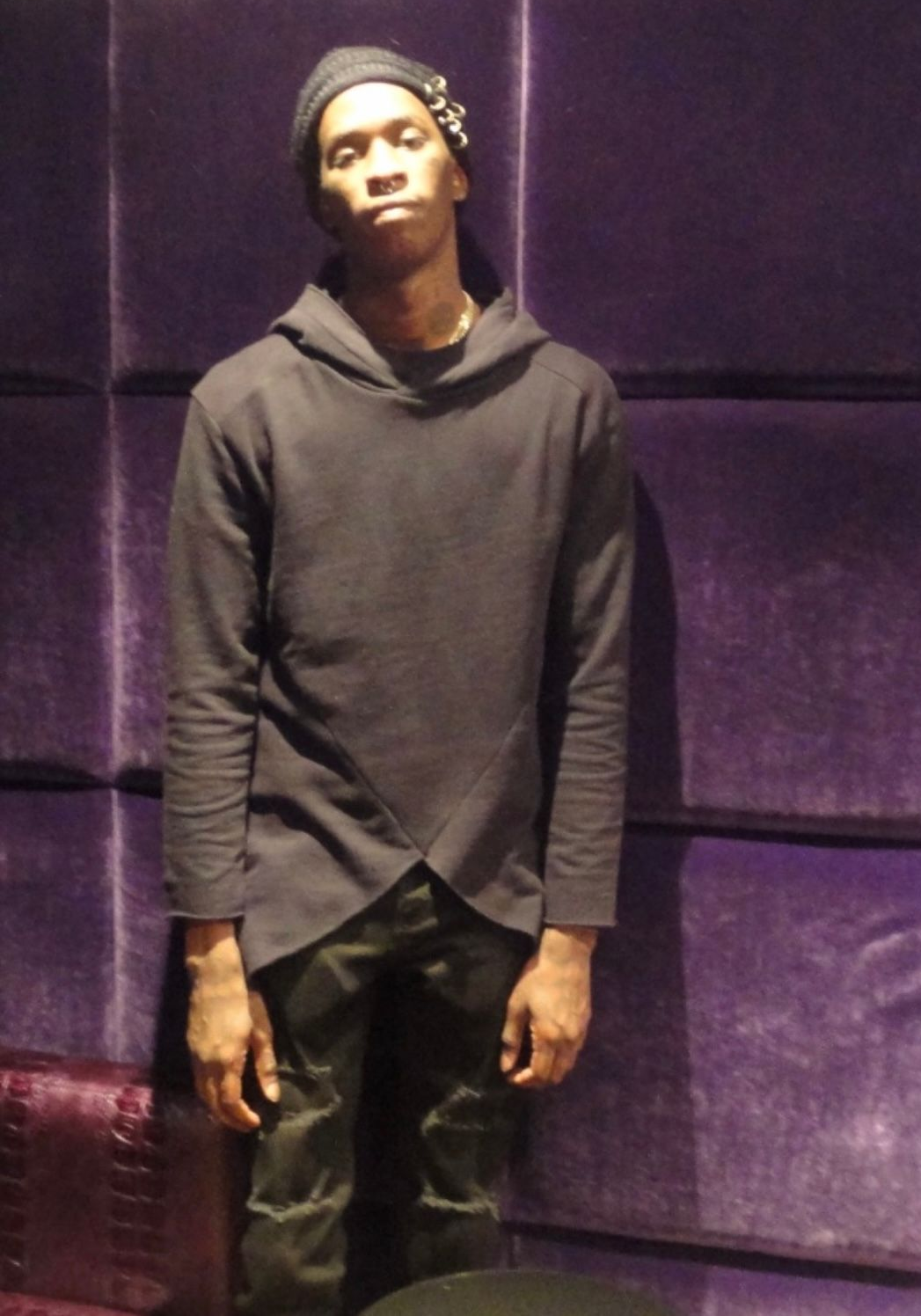A person in a dark hooded sweatshirt and beanie standing against a purple, padded wall.