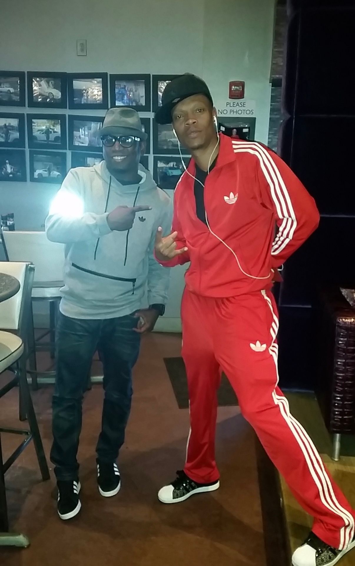 Two people pose in a room decorated with framed photos on the wall. One wears a grey hoodie, the other a red tracksuit.