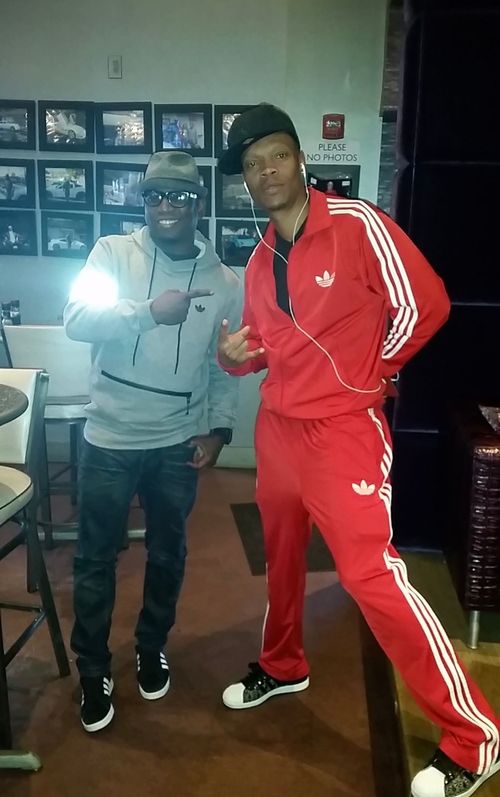 Two individuals stand in a room with framed photos on the wall; one wears a gray hoodie, the other a red Adidas tracksuit.