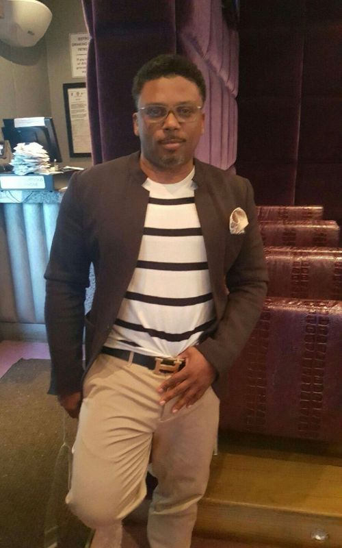 A person in a brown blazer, striped shirt, and tan trousers poses in a room with purple textured decor and wood stairs.