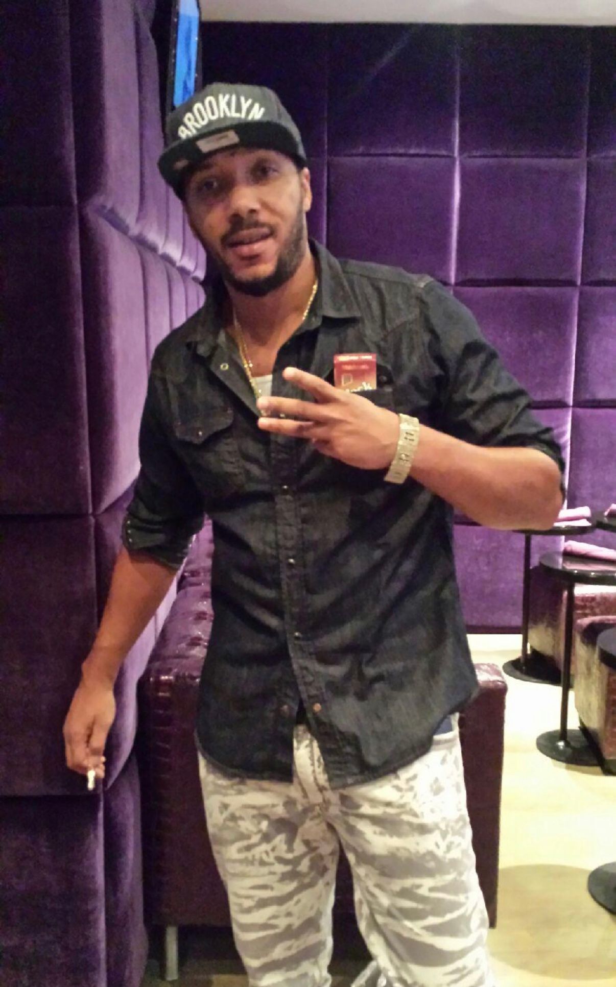 A person wearing a Brooklyn baseball cap and dark shirt poses in front of purple padded walls, making a hand gesture.