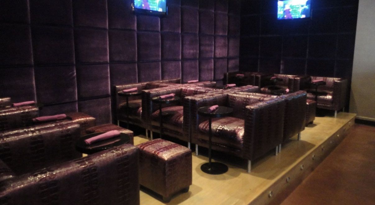 A lounge area features dark purple padded walls, tiered levels with leather seating, small side tables, and mounted TVs.