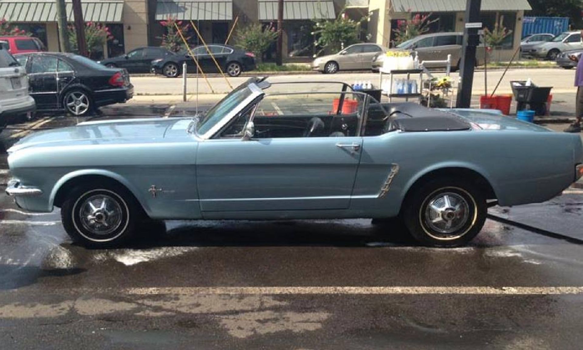 A light blue classic Ford Mustang convertible parked on a wet asphalt street in front of a row of storefronts.
