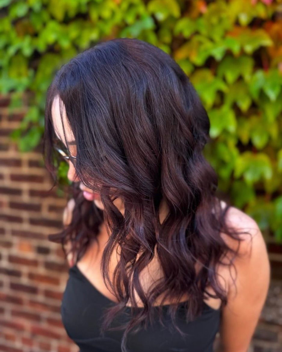A woman with long dark hair is standing in front of a brick wall.