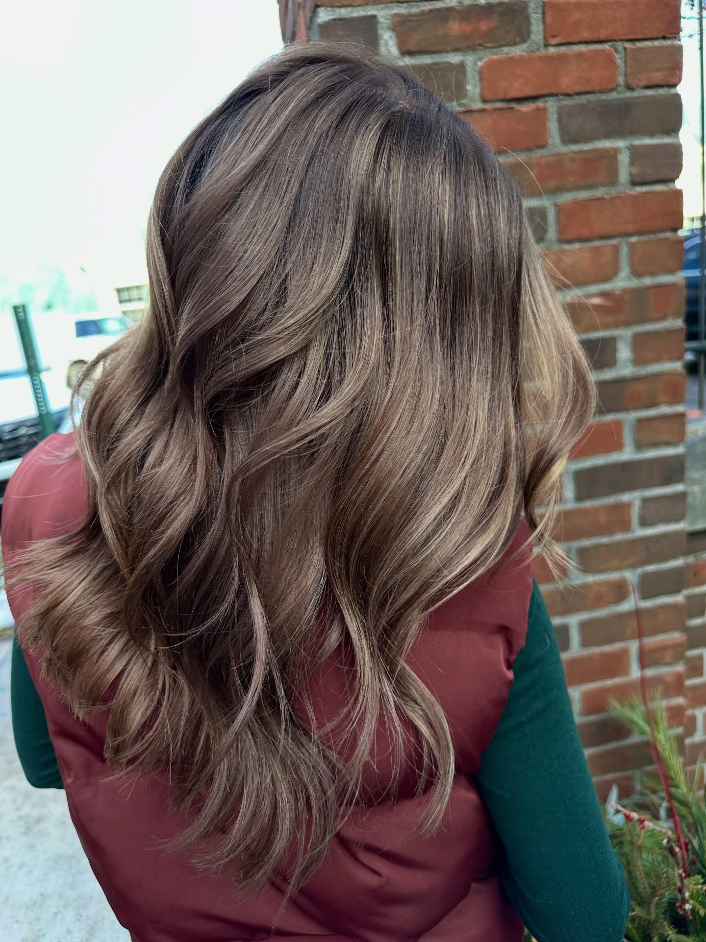 Woman with wavy, brown hair. Wearing a red vest and a green shirt. Near a brick wall.
