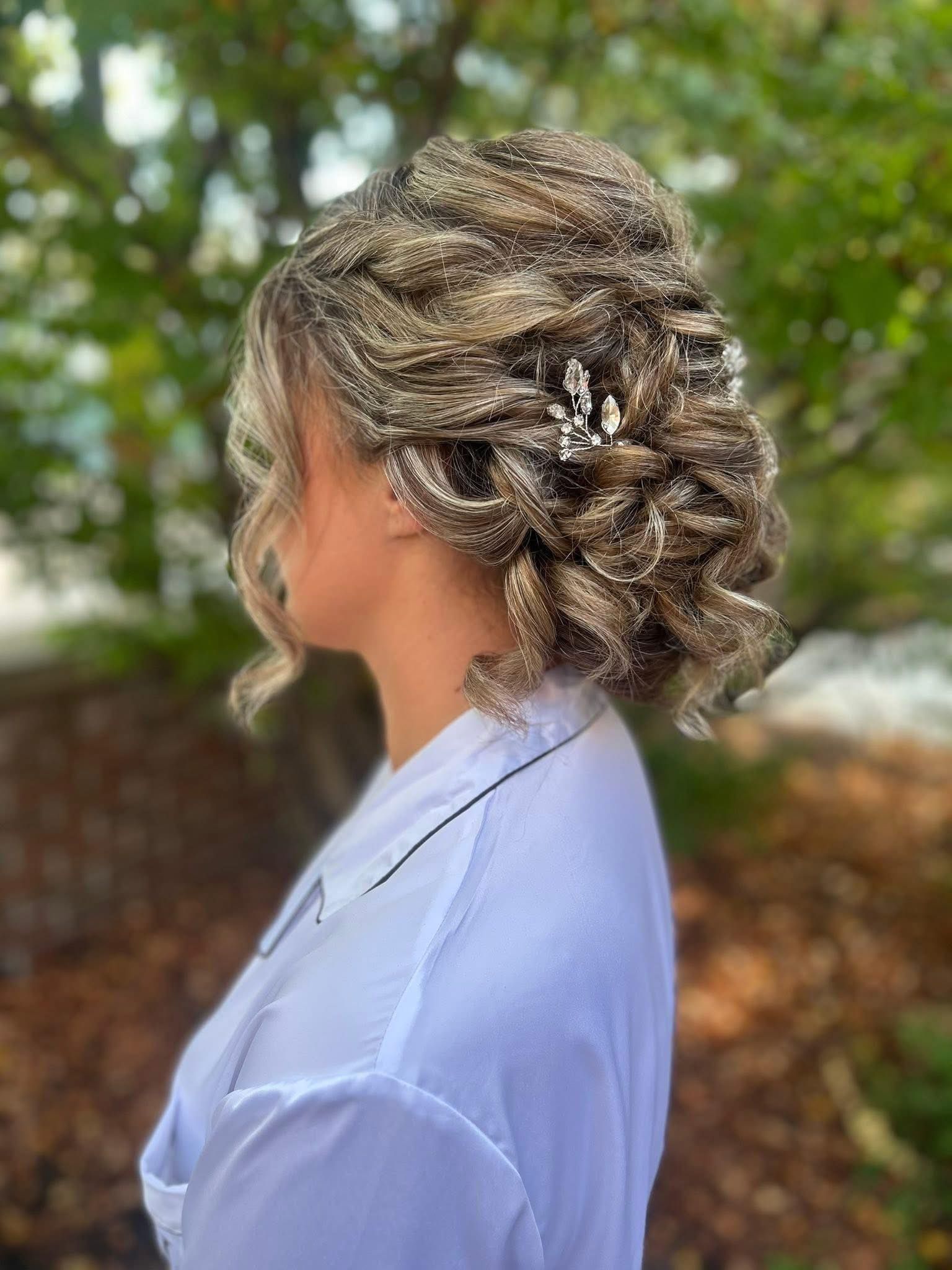 Woman with curled updo hairstyle, wearing a white top, set against a blurred green and brown background.