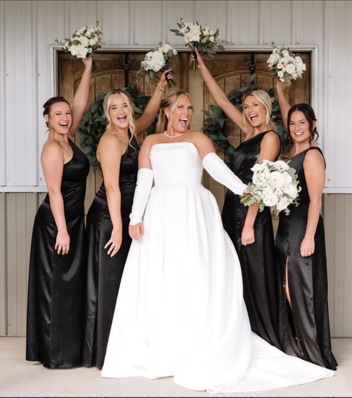 Bride with bridesmaids in black dresses, all holding bouquets and smiling outdoors.