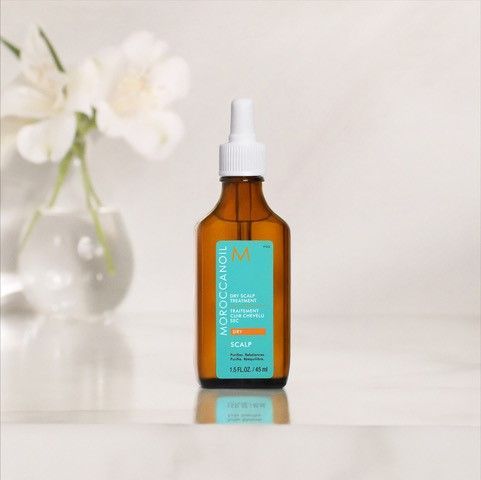 A bottle of Moroccan Oil hair oil is sitting on a table next to a vase of flowers.