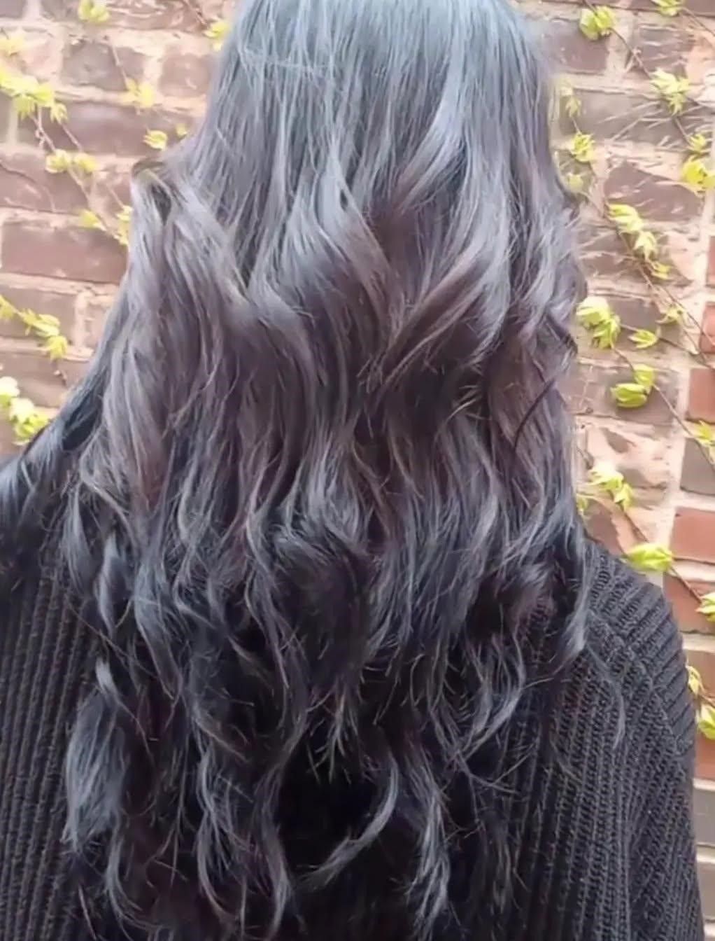 A woman with long black hair is wearing a black sweater and standing in front of a brick wall.