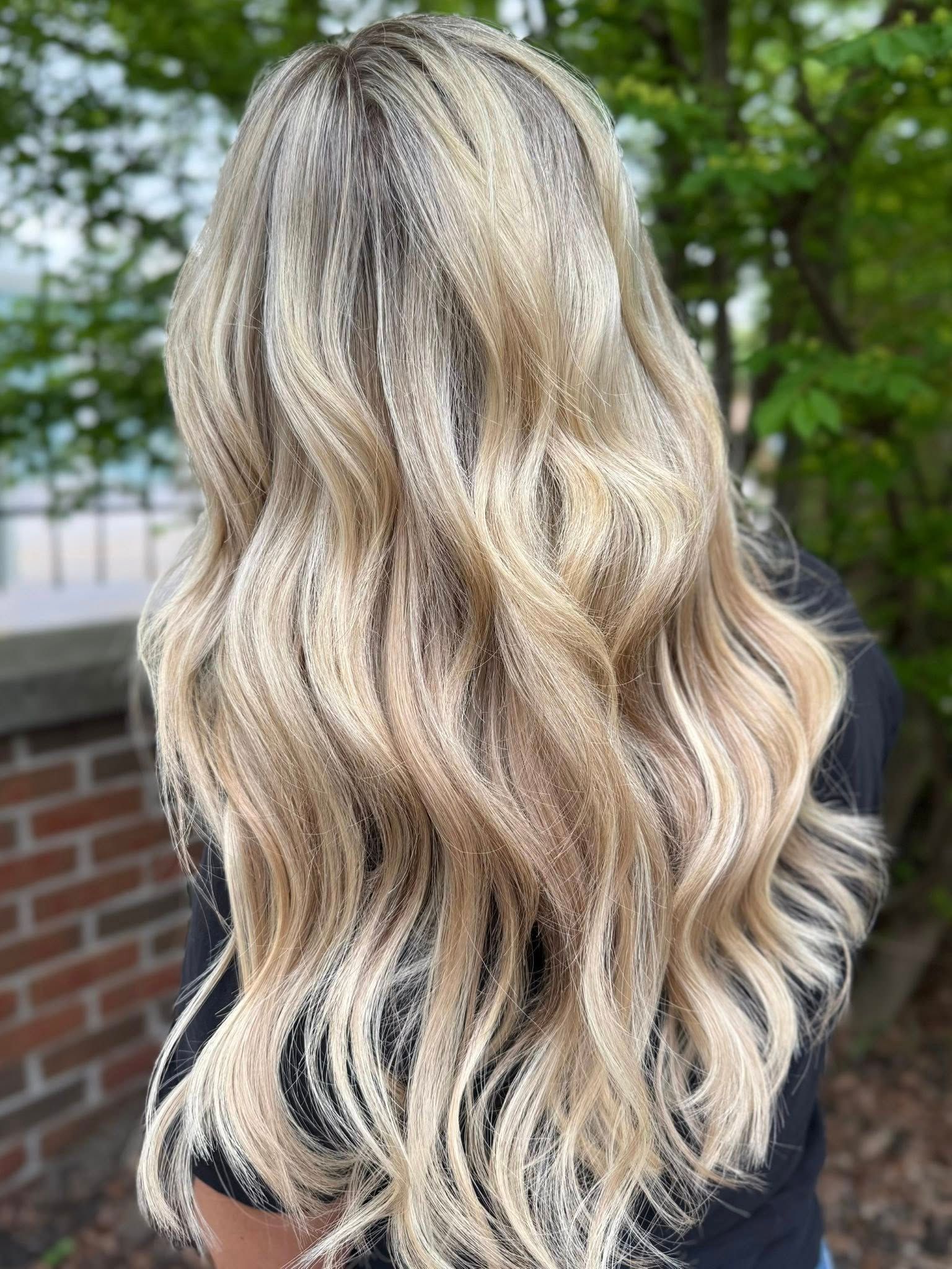 Long wavy blonde hair viewed from the back, outdoors near a brick wall and trees