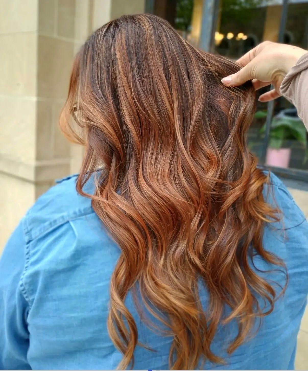Long wavy brown hair with copper highlights, styled in layers as a hand lifts a section outside a salon
