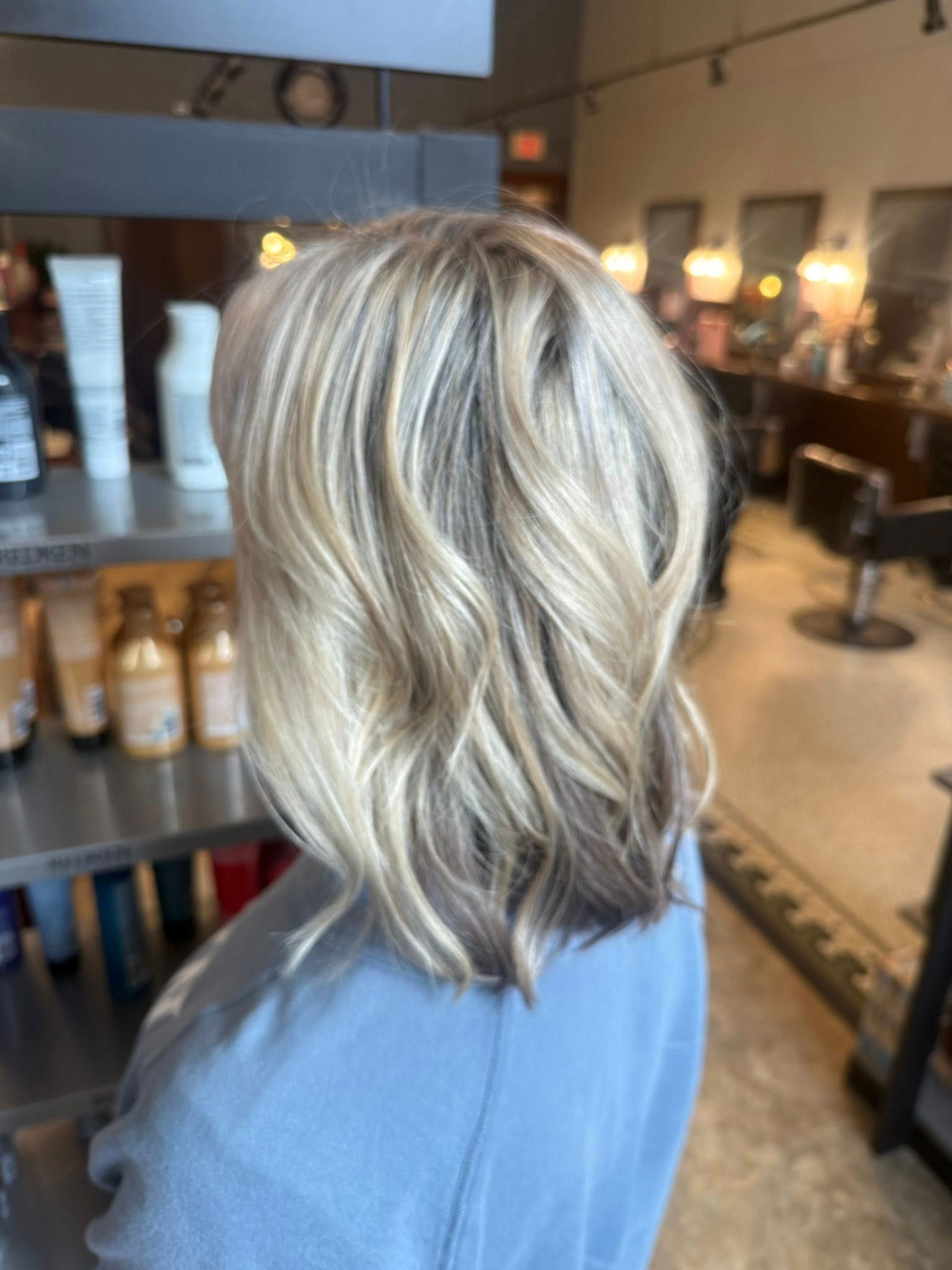 Short wavy blonde bob with dark roots, shown from the back in a salon.