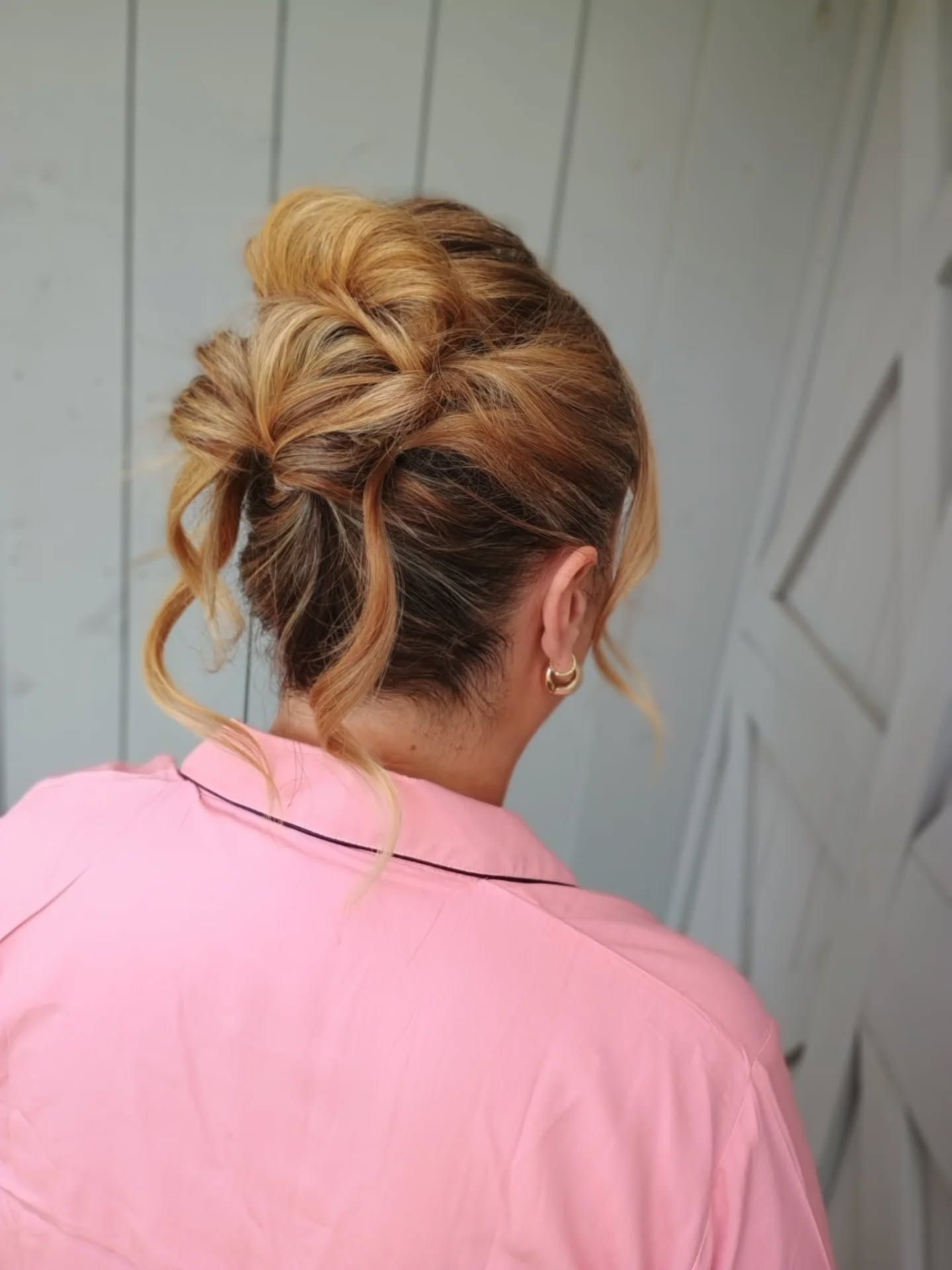 Back view of a blonde person with an elegant updo, wearing a pink shirt against a gray wall.