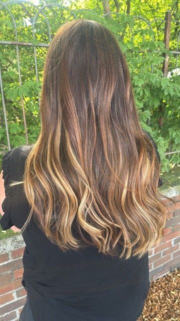 Back view of long wavy brown hair with blonde highlights, standing outdoors by greenery and a brick wall