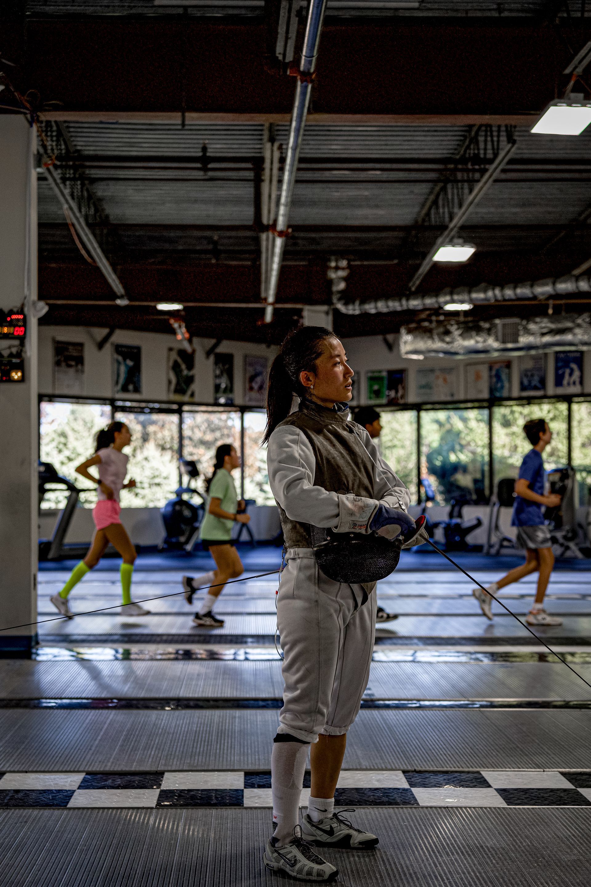 A woman is standing in a gym wearing a fencing outfit.