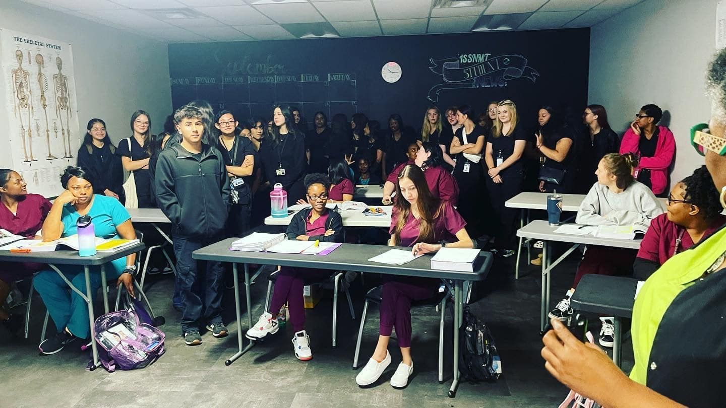 Classroom of medical students in scrubs, some seated at desks, others standing. A black chalkboard in the background.