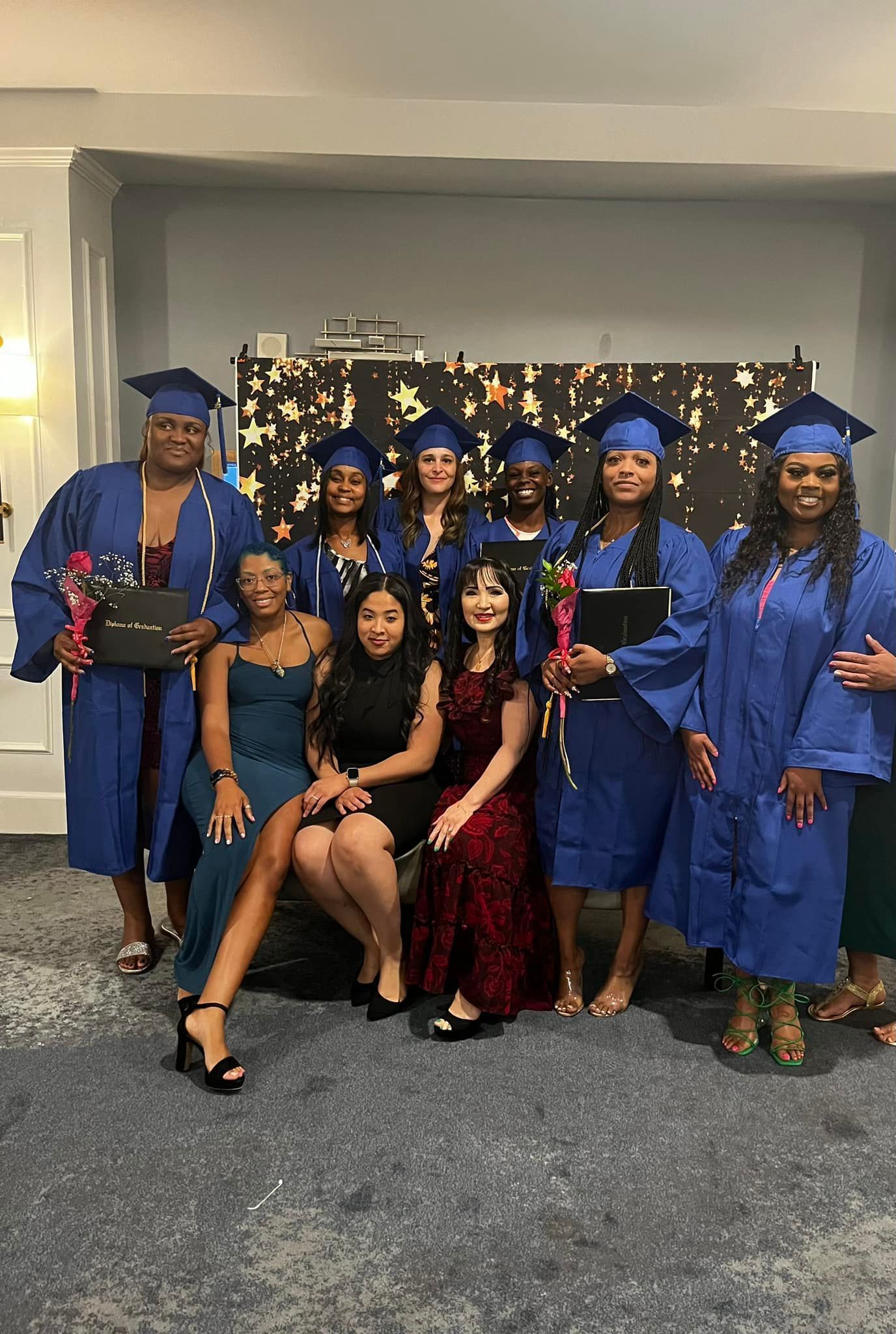 Group of graduates in blue gowns and caps pose with friends in a celebratory room.