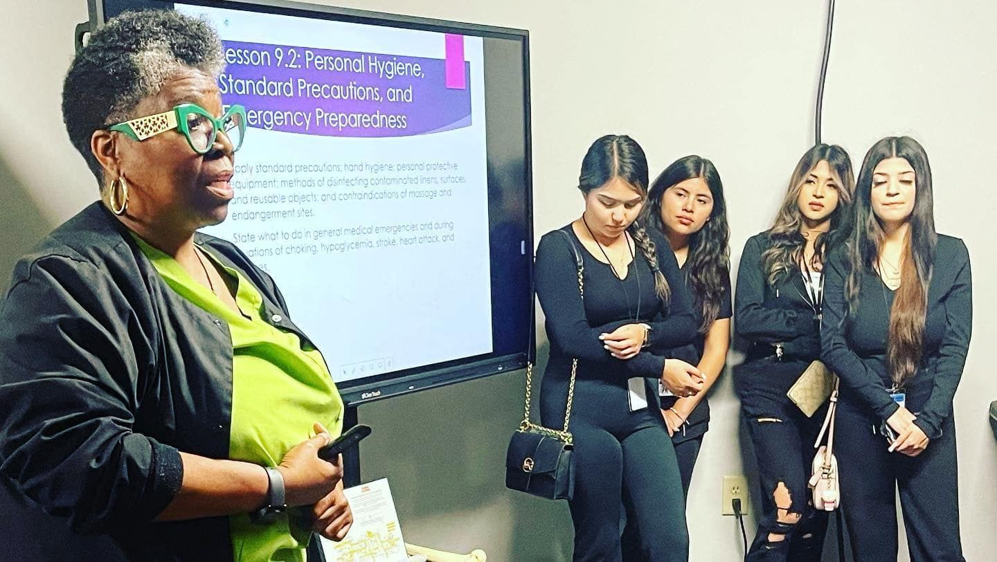 A woman in glasses presents, looking at a screen with text, while several women listen, in an office.