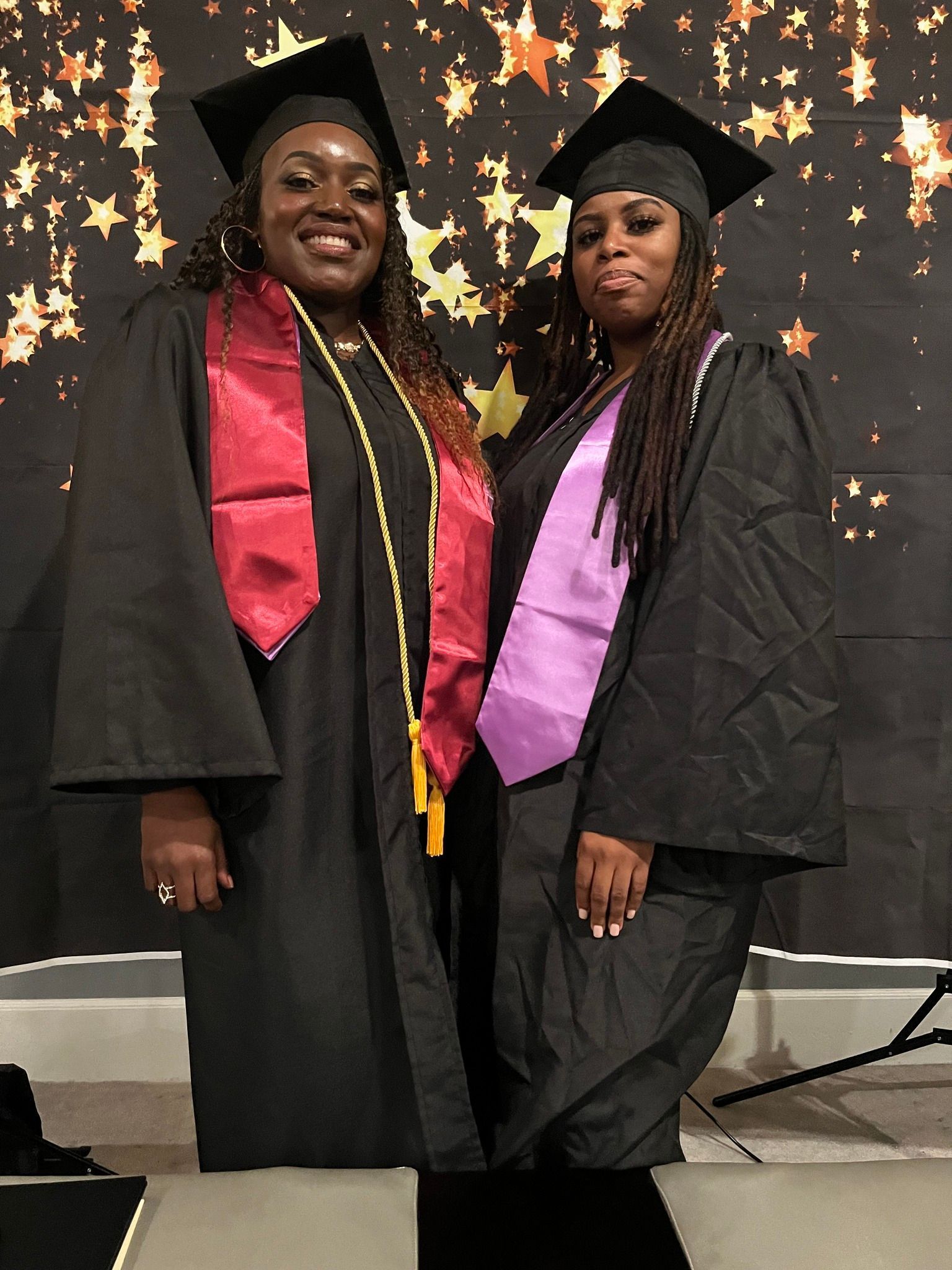 Two people in graduation gowns, smiling. One wears a red sash, the other purple, in front of a star backdrop.