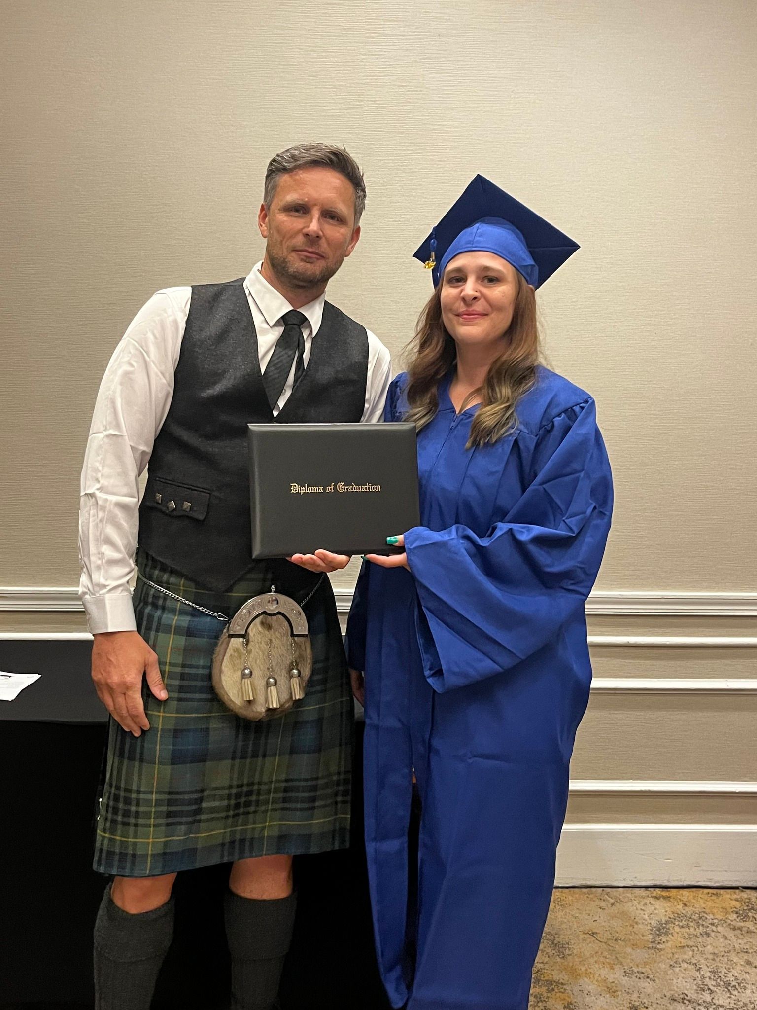 Man in kilt and woman in graduation gown holding a diploma in a room, both smiling.