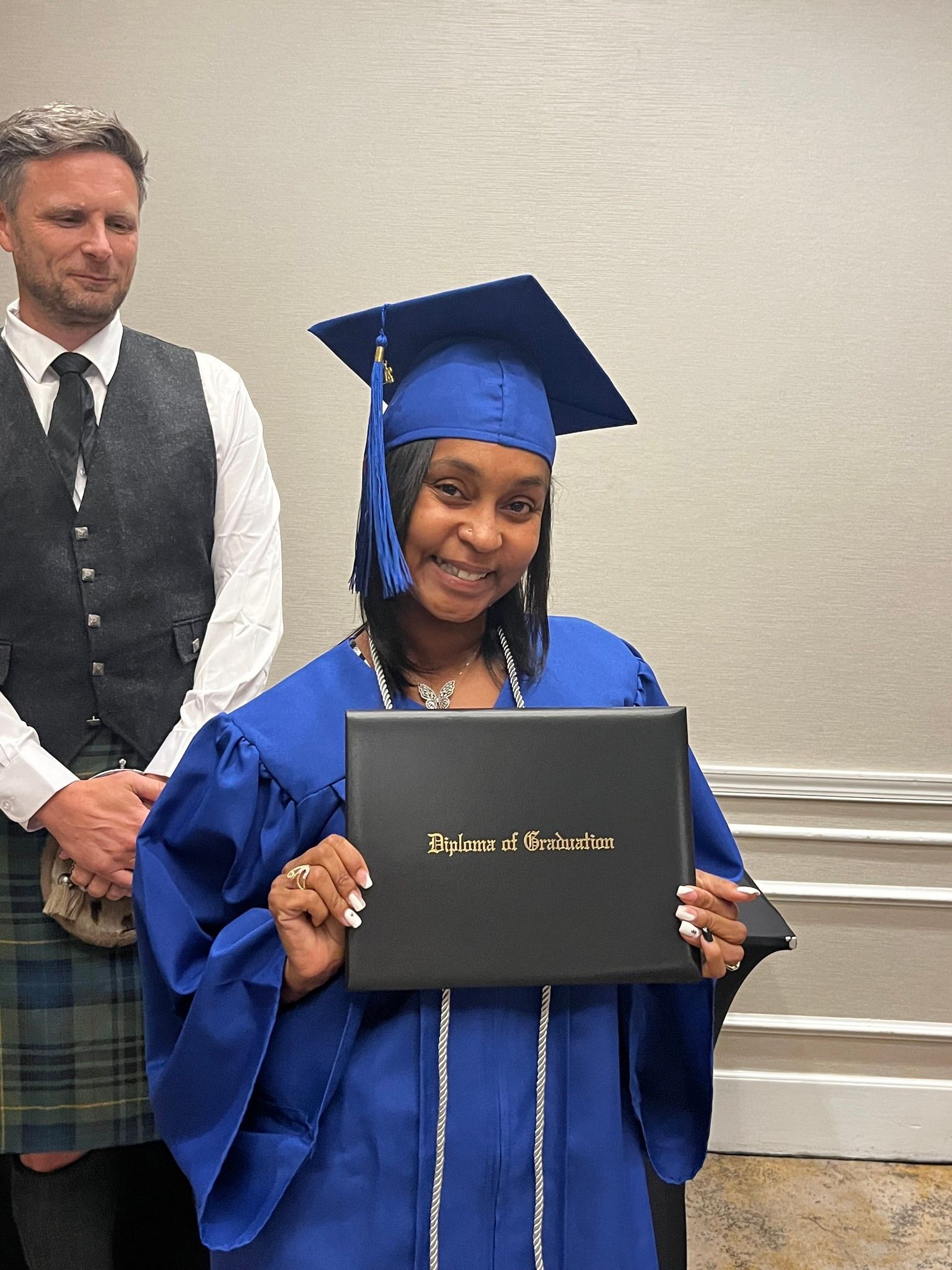 Person in blue graduation gown holding diploma, smiling. Man in kilt stands behind.