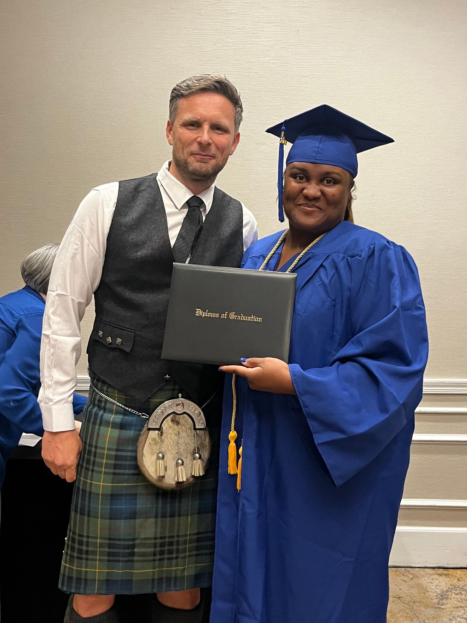 Man in kilt and woman in graduation gown hold a diploma. They smile, in a room with others in blue.