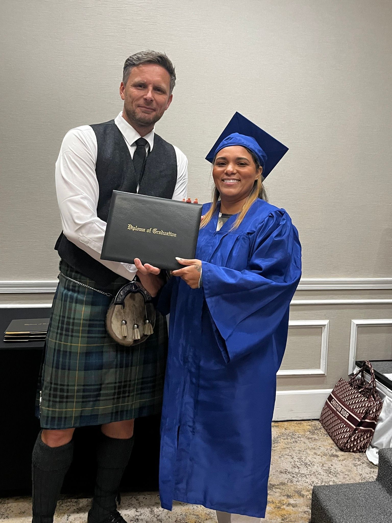 Man in kilt presents a diploma to a woman in a blue graduation gown. They are smiling.