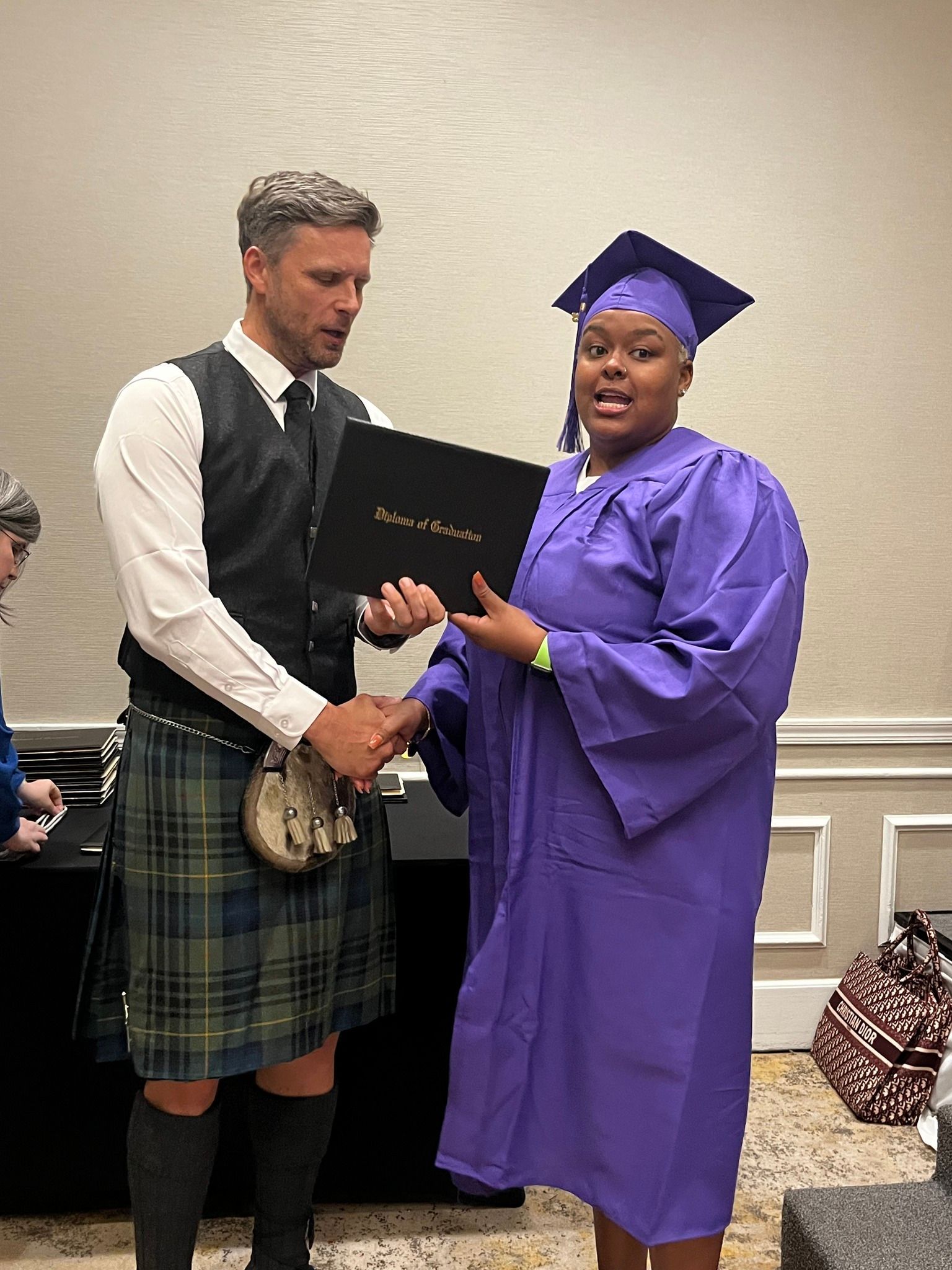 Man in kilt hands a diploma to person in purple graduation gown and cap.