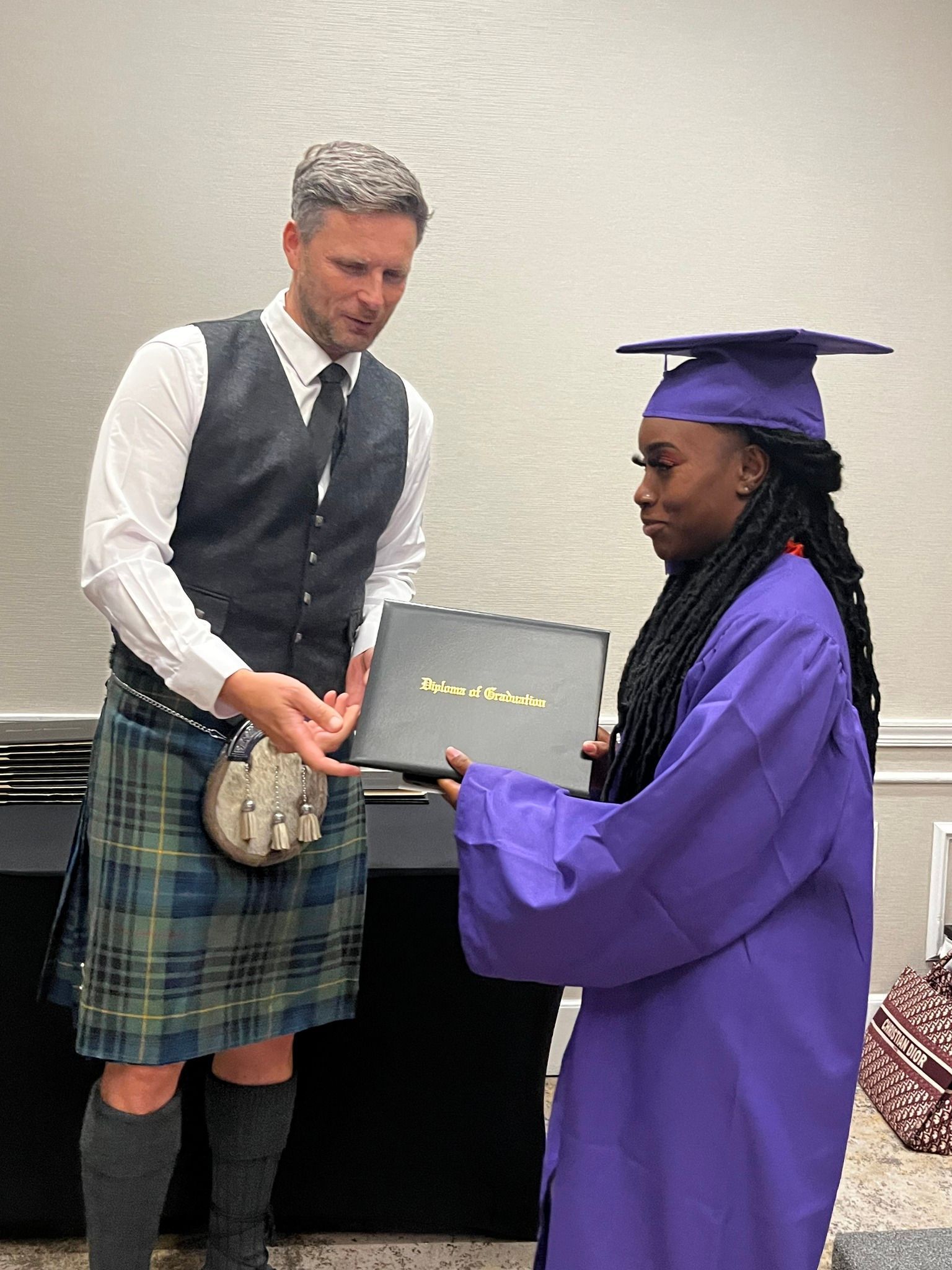 Man in kilt hands diploma to person in purple graduation gown. Indoor setting.