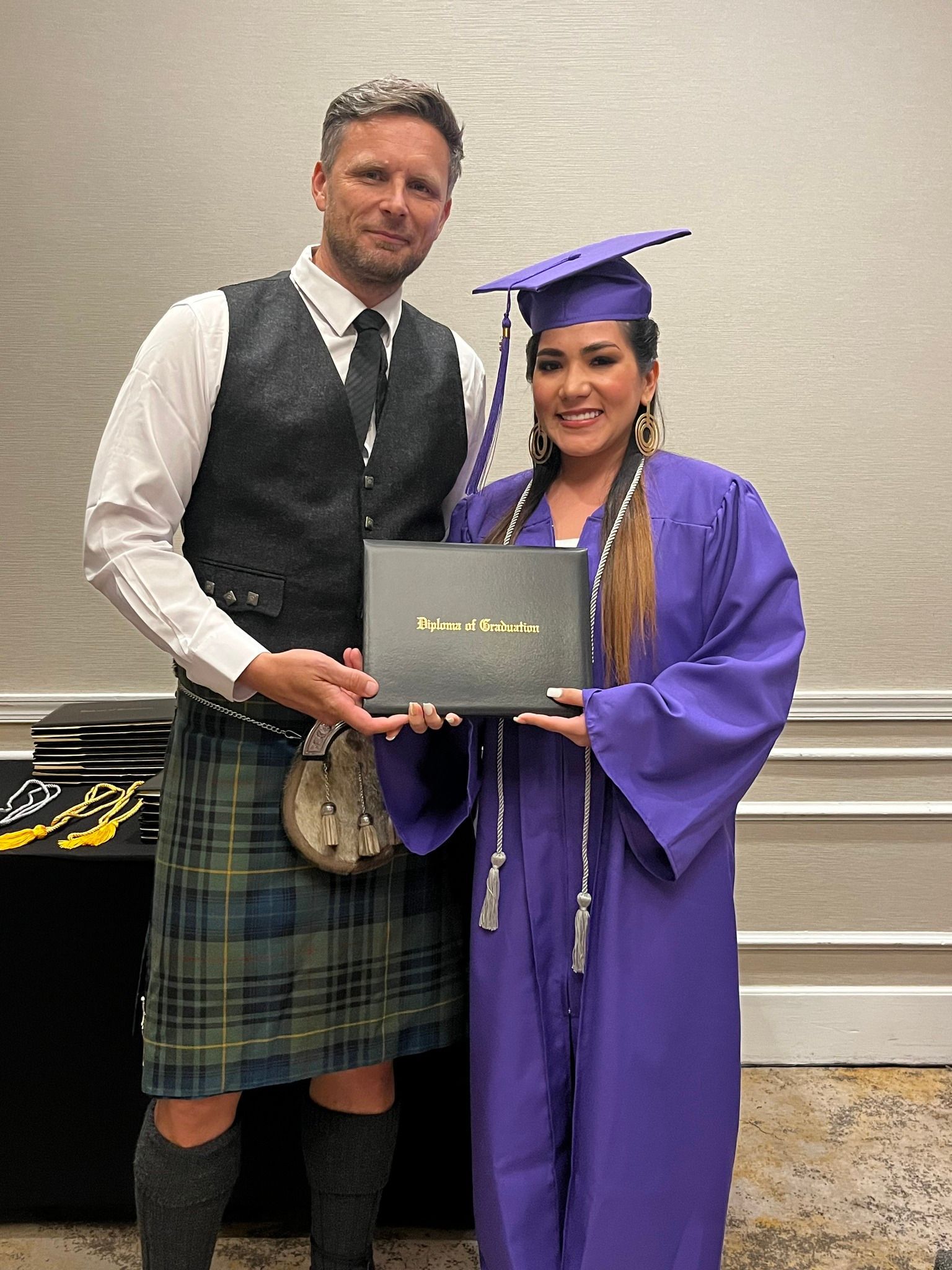 Woman in purple graduation gown and man in kilt hold diploma; setting is an event.