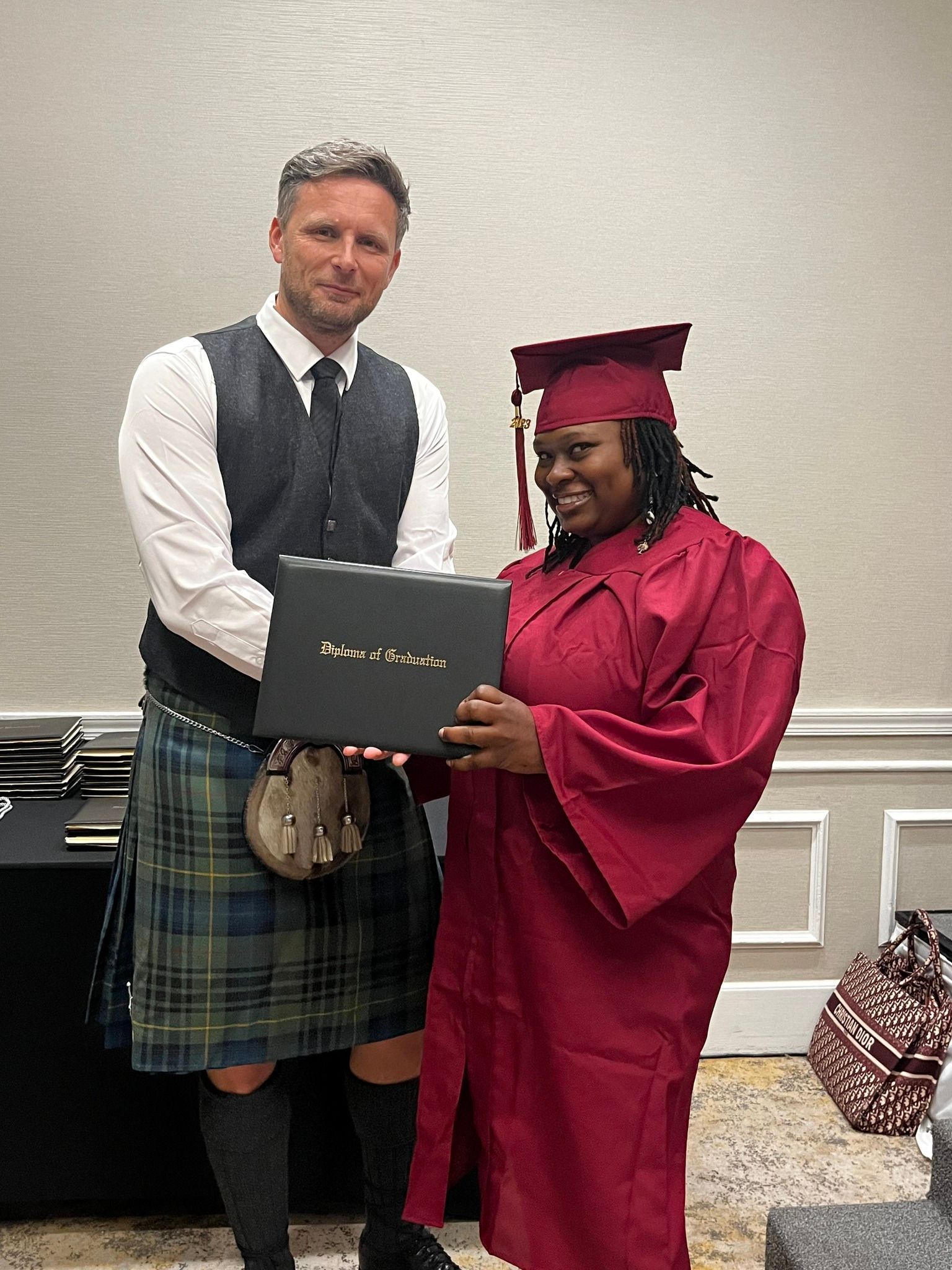 Man in kilt hands diploma to person in burgundy graduation gown. Hotel setting.