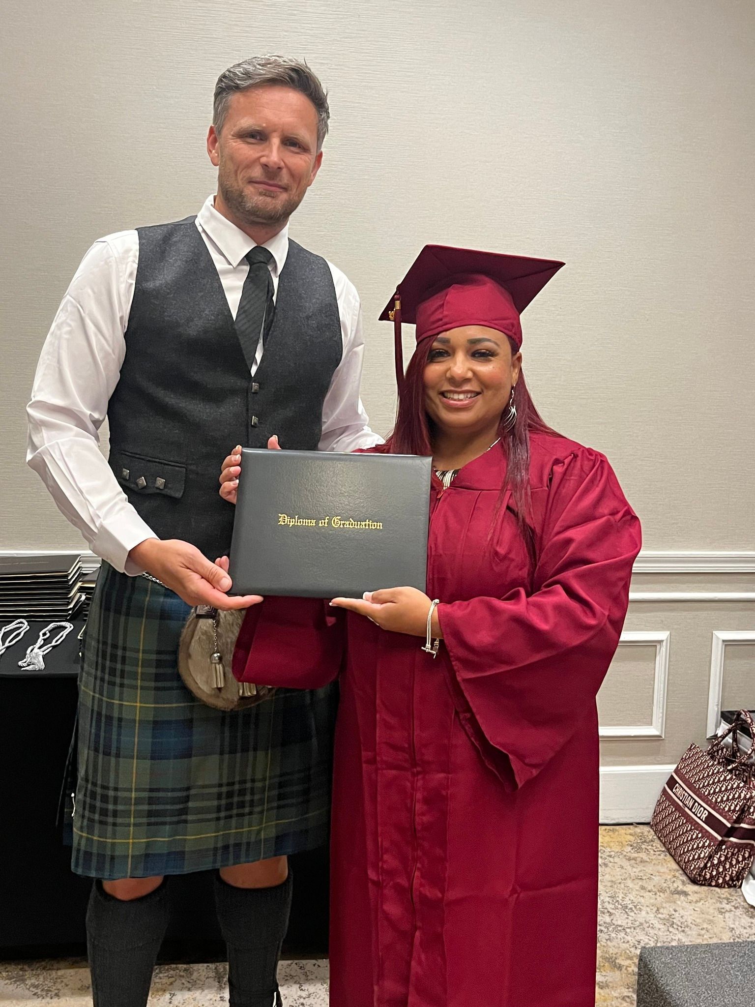 Man in kilt and woman in graduation robe holding diploma. Formal setting, smiling.