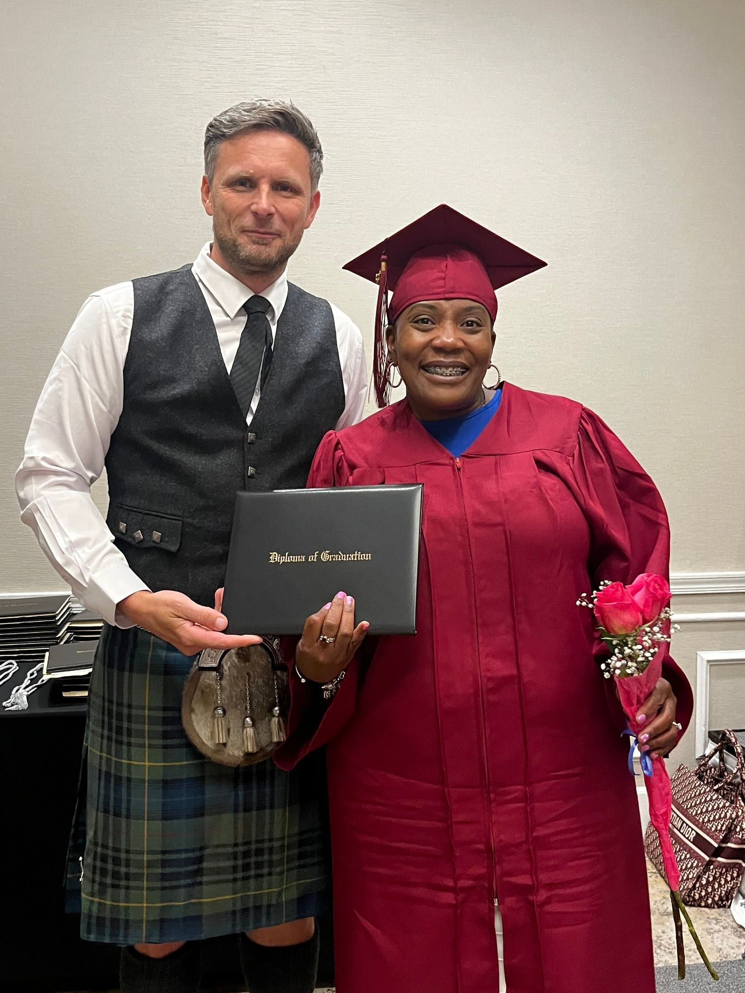 Man in kilt and woman in graduation gown holding a diploma and rose, indoors.