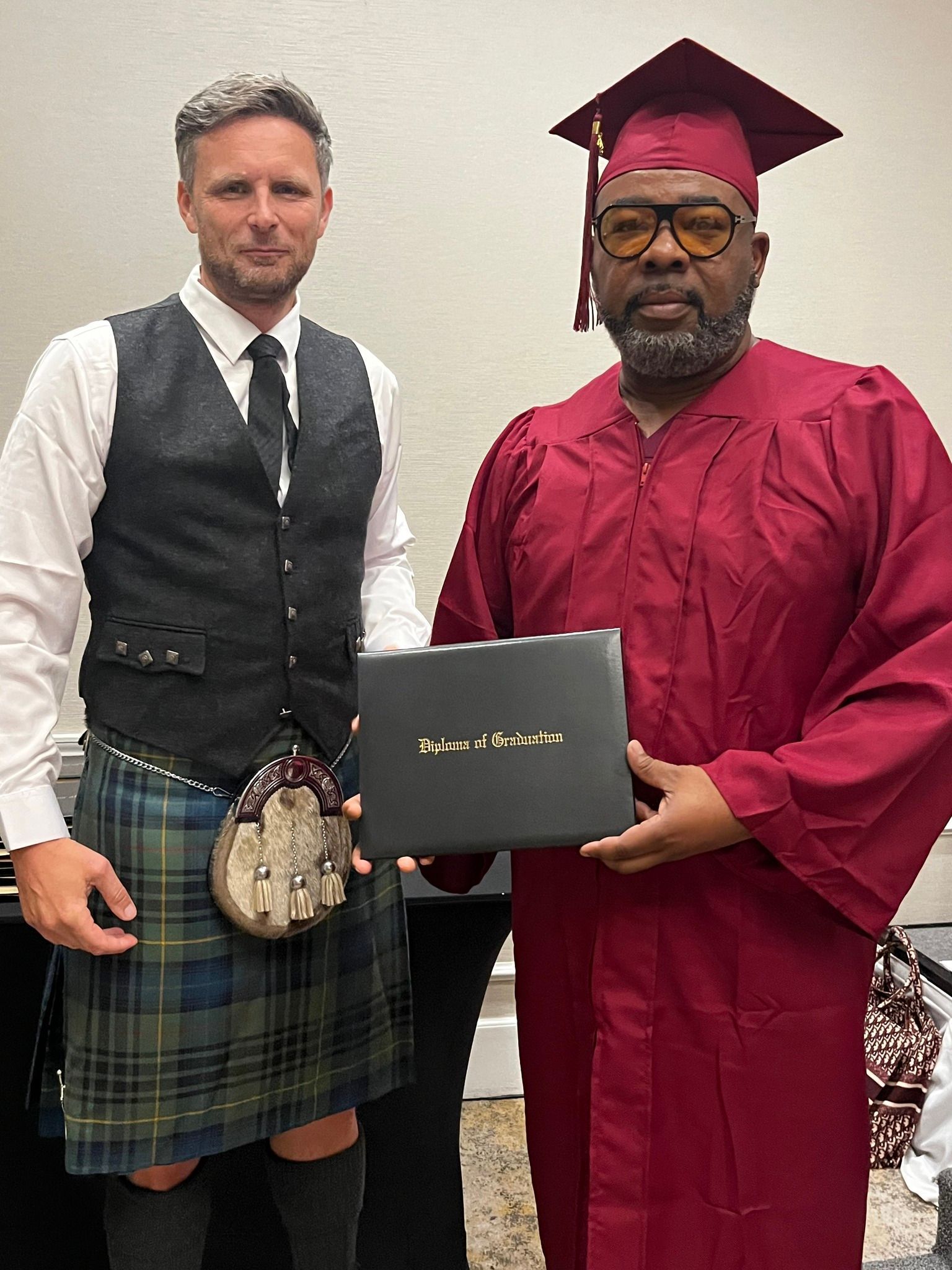 Man in graduation gown and man in kilt holding a diploma. They are inside a room with a cream wall.