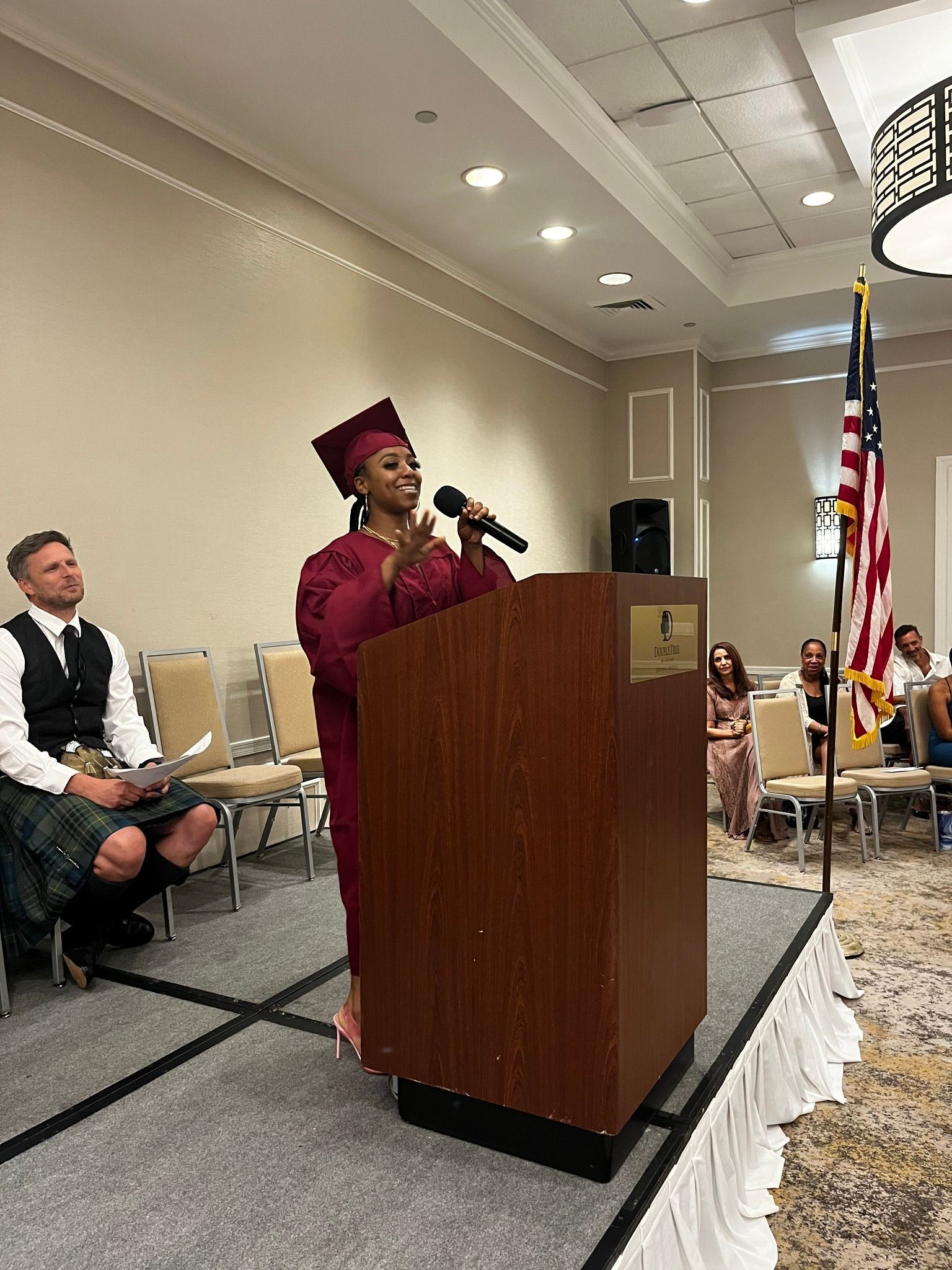 A person in maroon graduation attire speaks at a podium. Others are seated or standing in a hotel ballroom.